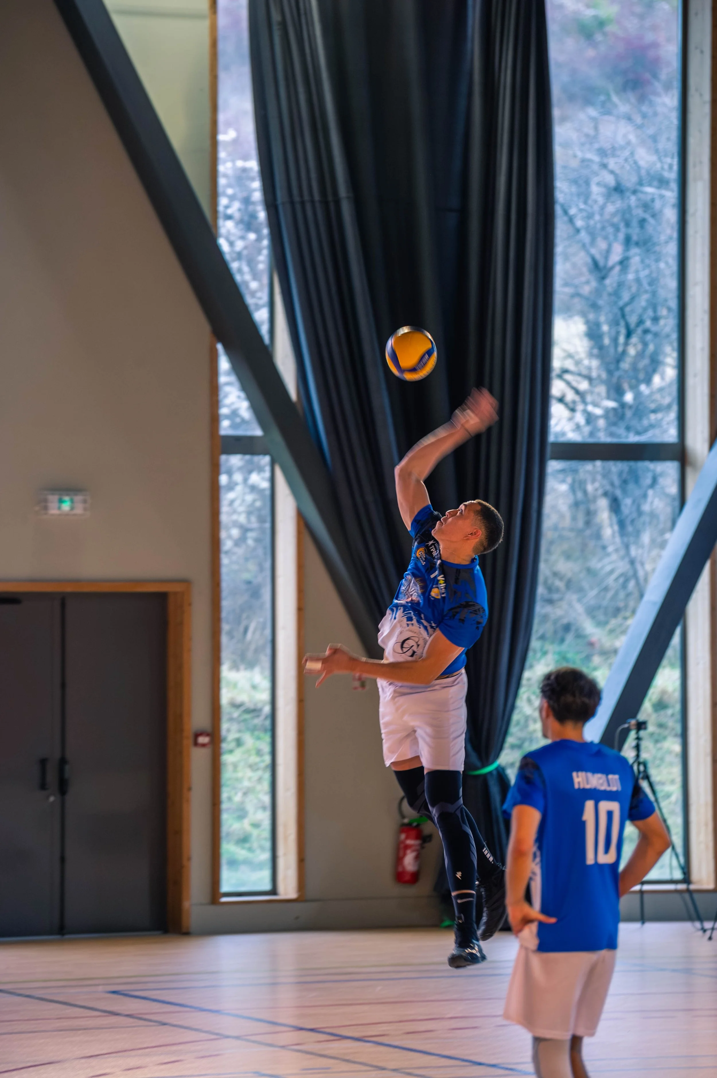 Deux jeunes hommes jouent au volleyball en intérieur. L'un d'eux, en plein saut, essaie de frapper le ballon, tandis que l'autre, portant un maillot avec le nom 'HUBERT' et le numéro 10, regarde le jeu. Le décor indique une salle avec de grandes fenê