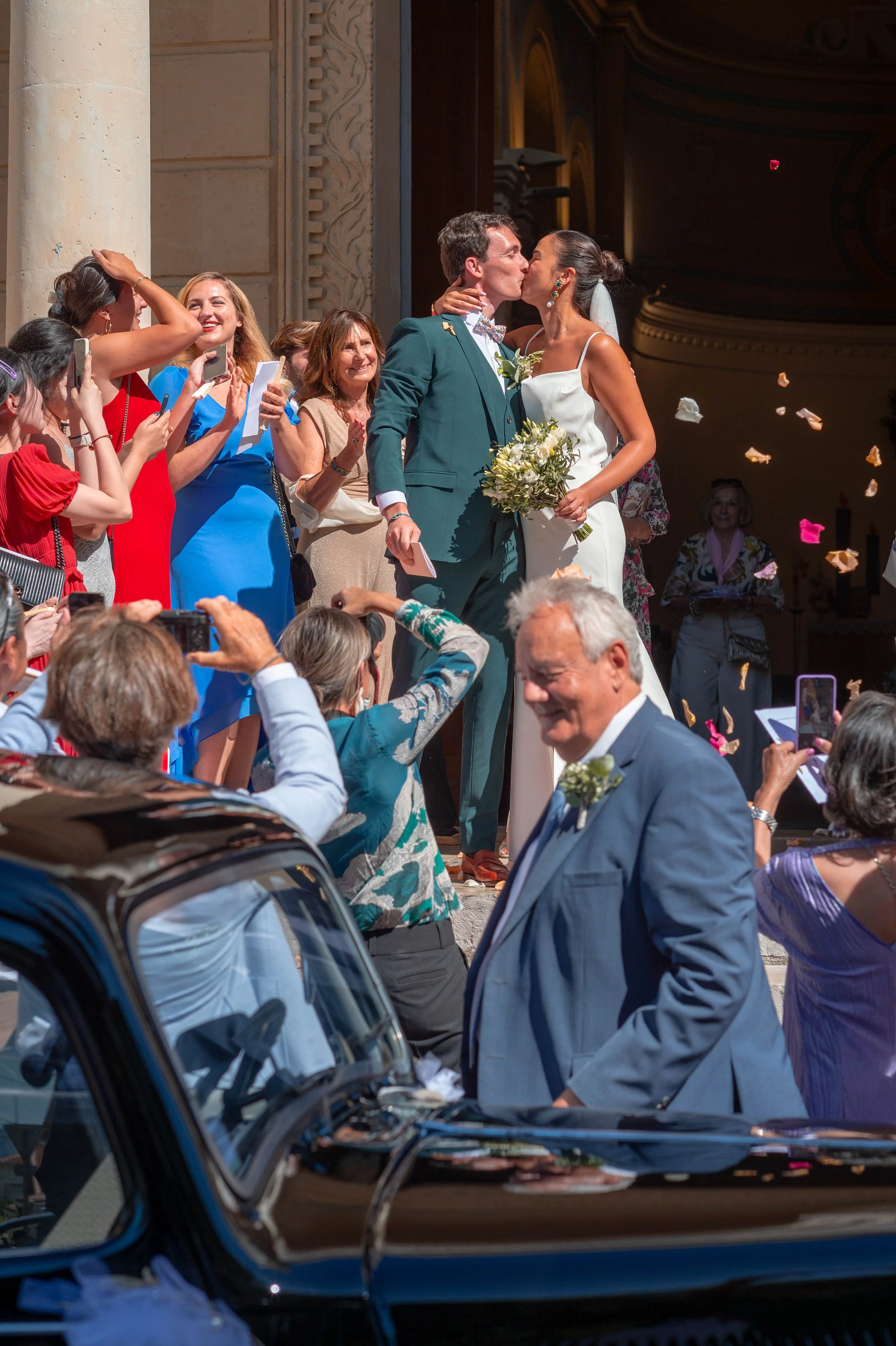 Un couple de mariés s'embrassent lors de leur mariage, entourés d'invités enchantés qui prennent des photos, avec des fleurs et des confettis en l'air.