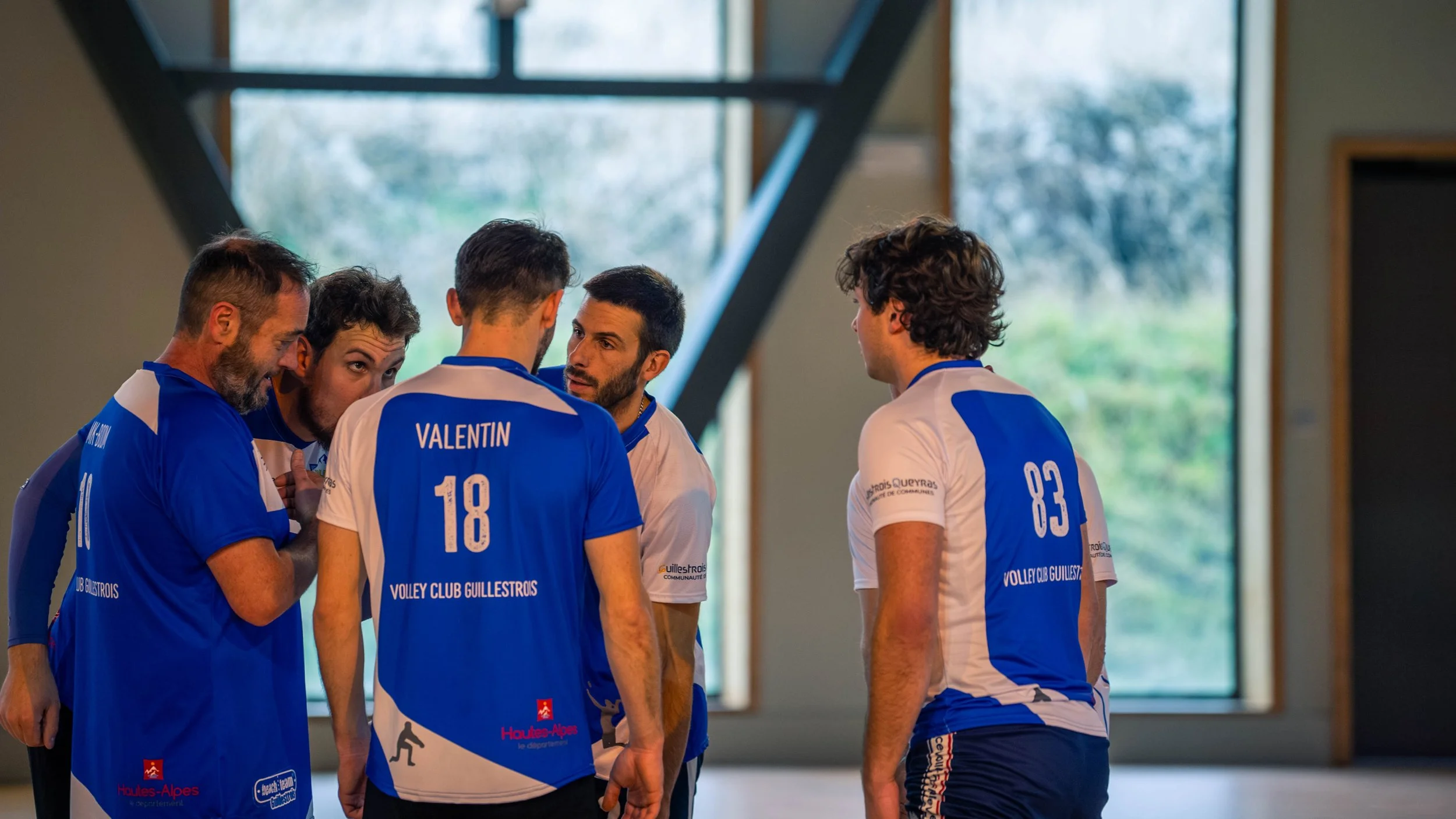 Cinq joueurs de volleyball du club Guilestroits en train de discuter dans un gymnase, portant des maillots bleus et blancs avec les numéros 18 et 83.