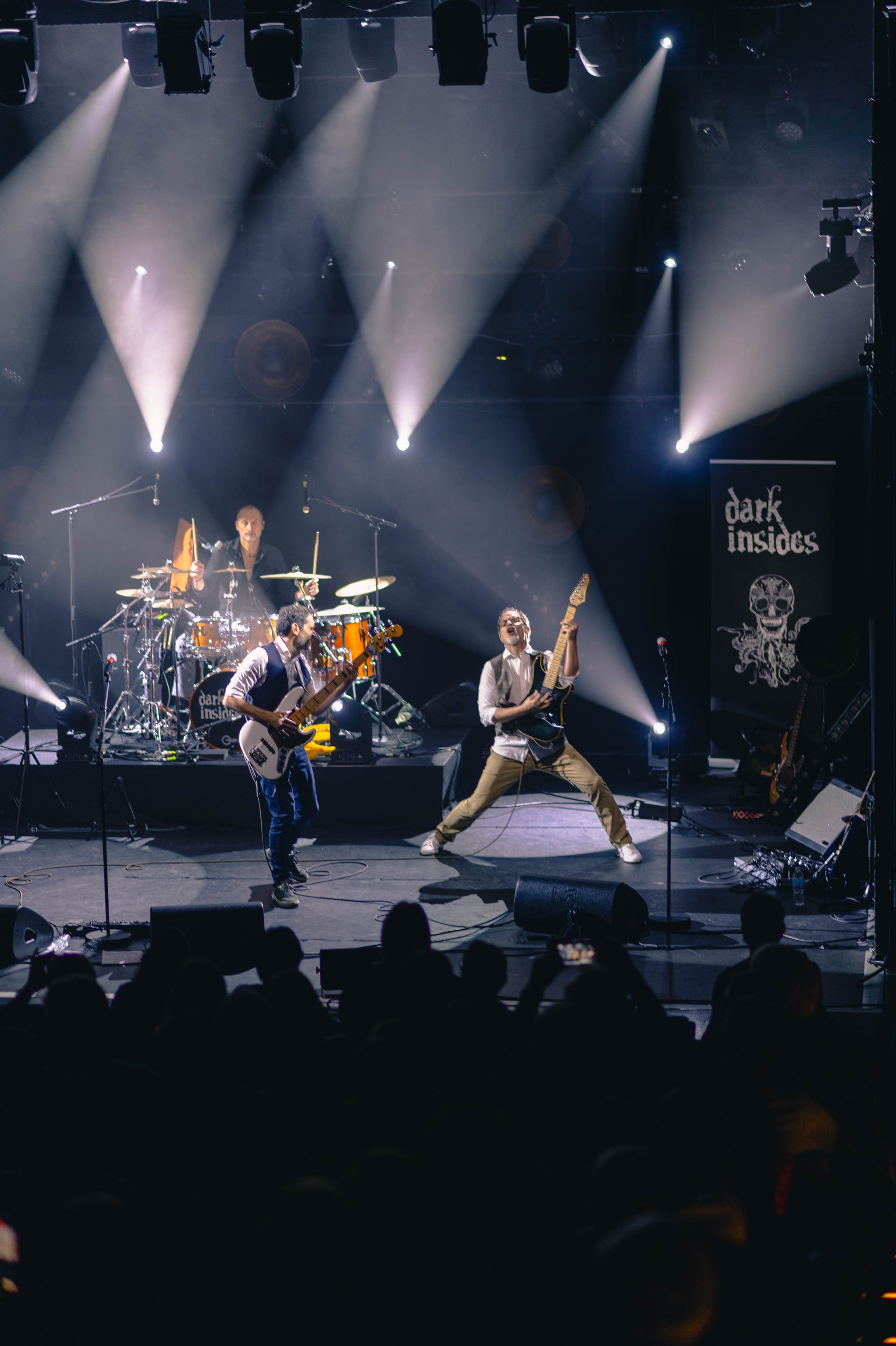 Un groupe de musiciens performe en direct sur scène avec des éclairages spectaculaires, incluant une batterie, deux guitares et un bassiste dans une salle de concert.