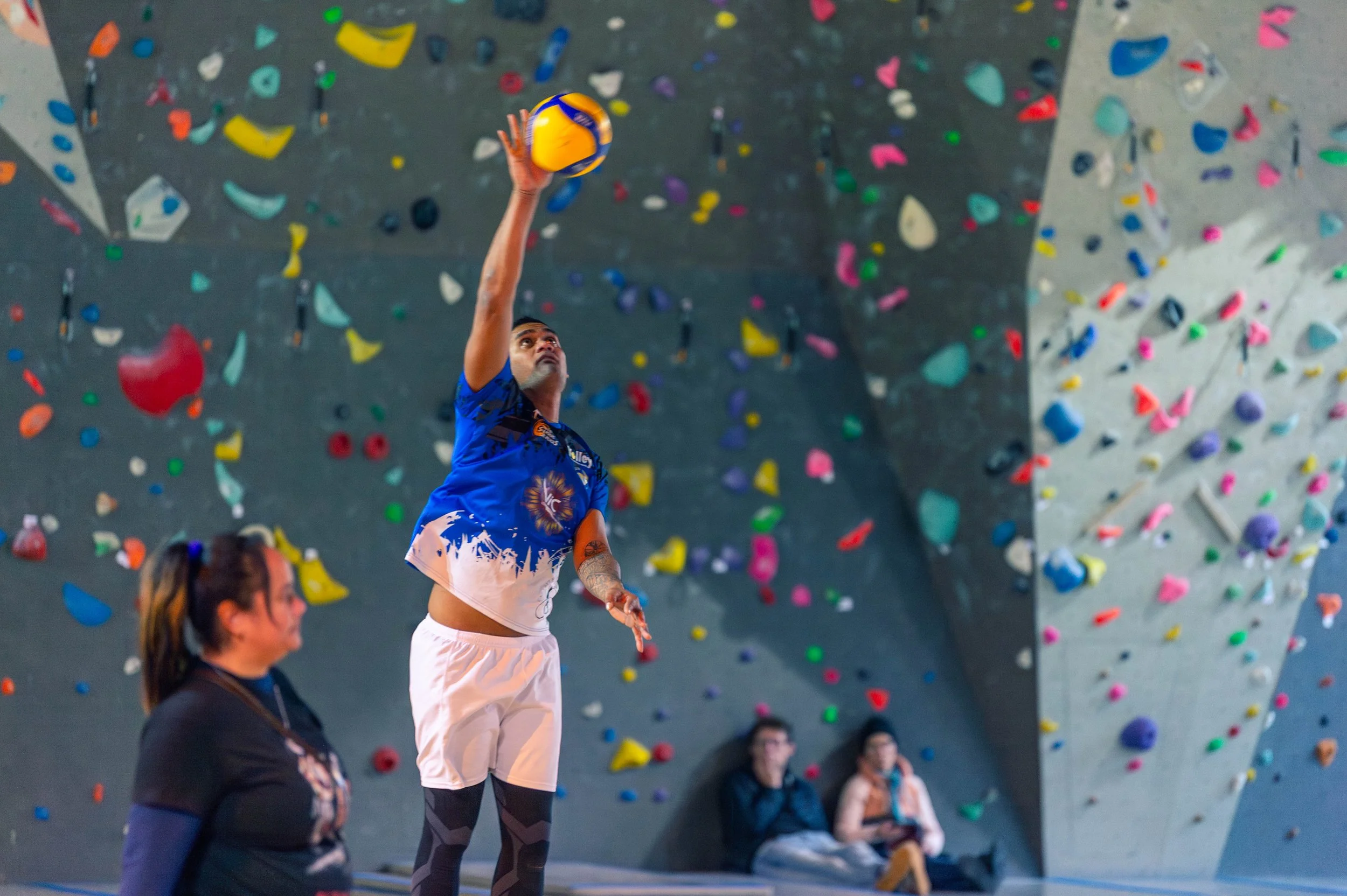 Un homme pratique le volleyball en salle, sautant pour frapper le ballon orange et jaune, avec un mur d'escalade coloré en arrière-plan et deux spectateurs assis observant la scène.