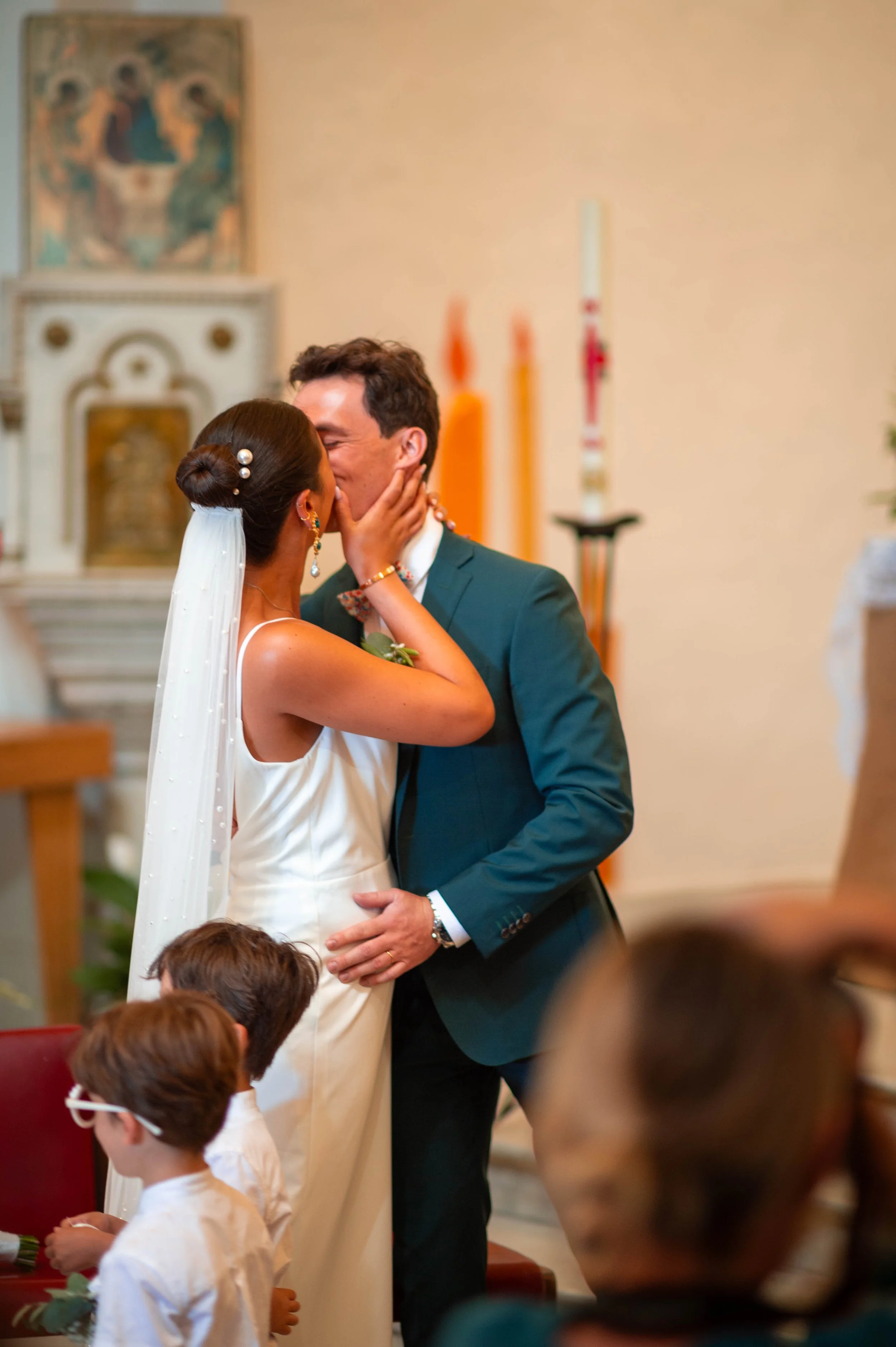 Un couple de mariés s'embrassant lors de leur mariage religieux dans une église, avec des enfants en premier plan et des bougies en arrière-plan.