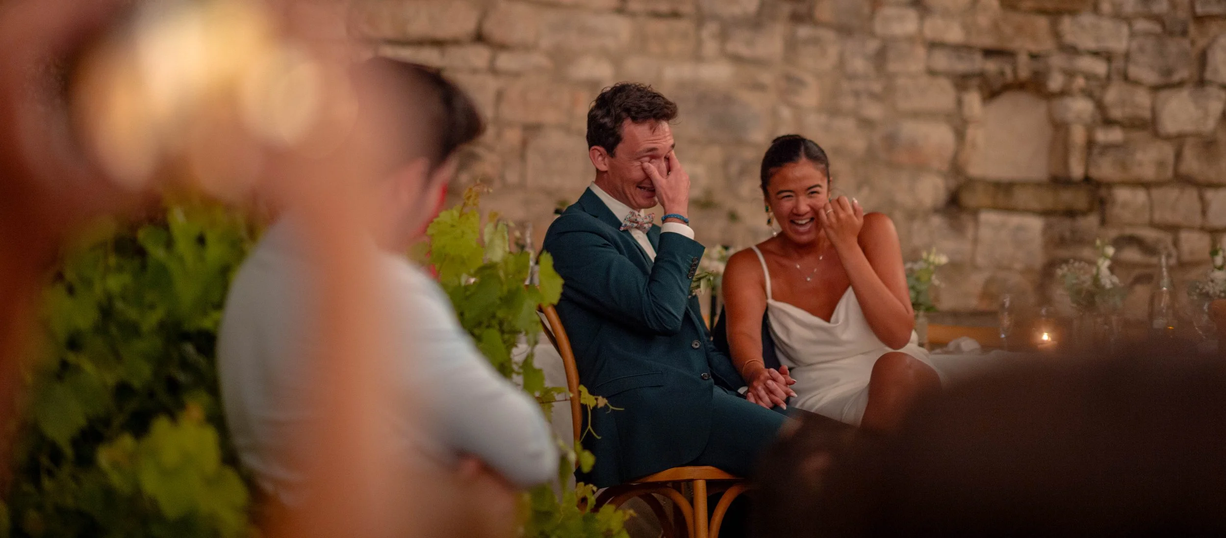 Deux personnes assises, riant et amusing à un événement, avec un mur en pierre en arrière-plan, décorations florales et bougies autour.