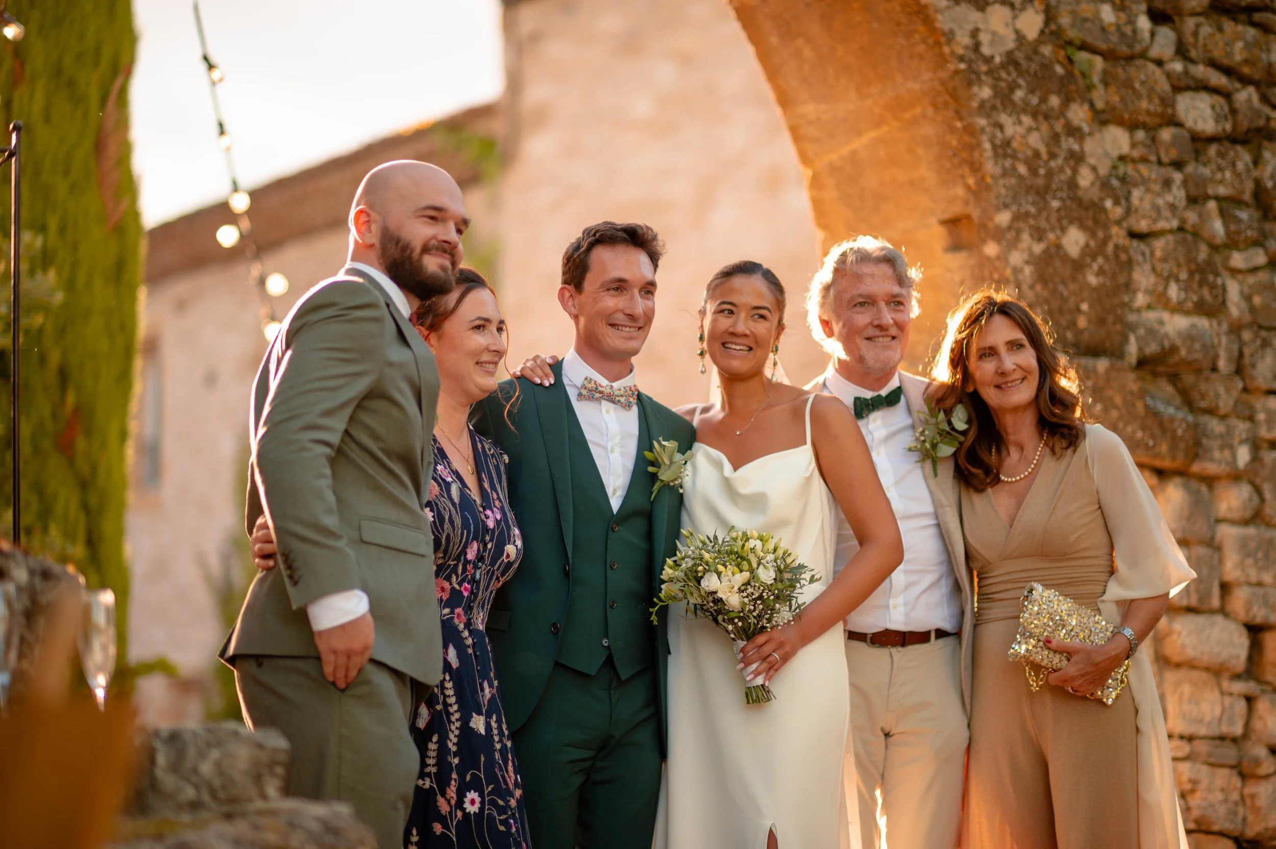 Groupe de six personnes souriantes lors d'un mariage en plein air, ambiance chaleureuse avec coucher de soleil, devant un mur en pierre.