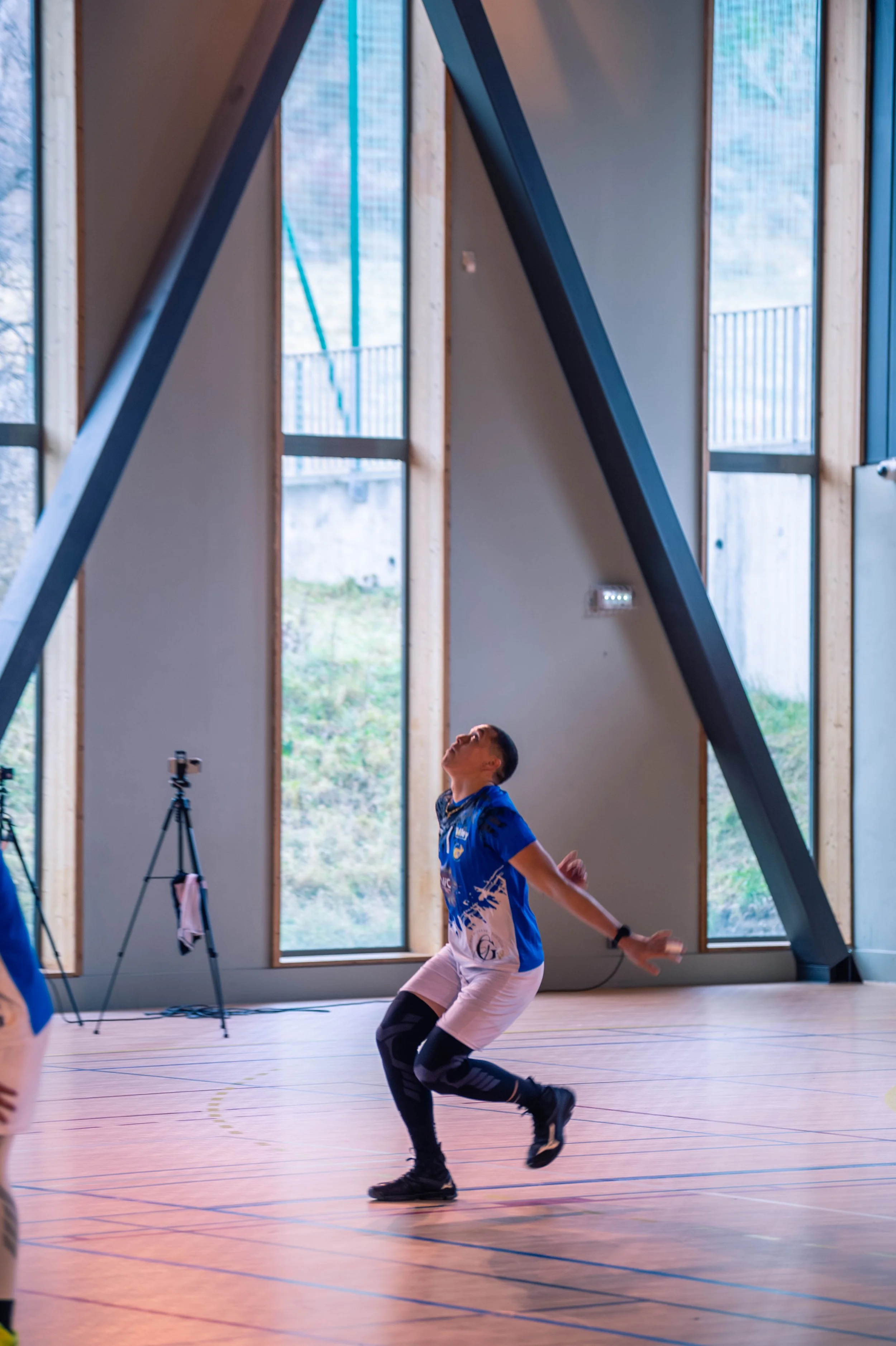 Un joueur de volleyball en intérieur, portant un maillot bleu et un équipement de genou, dans une posture prête à faire un mouvement lors d'un match ou d'une pratique.