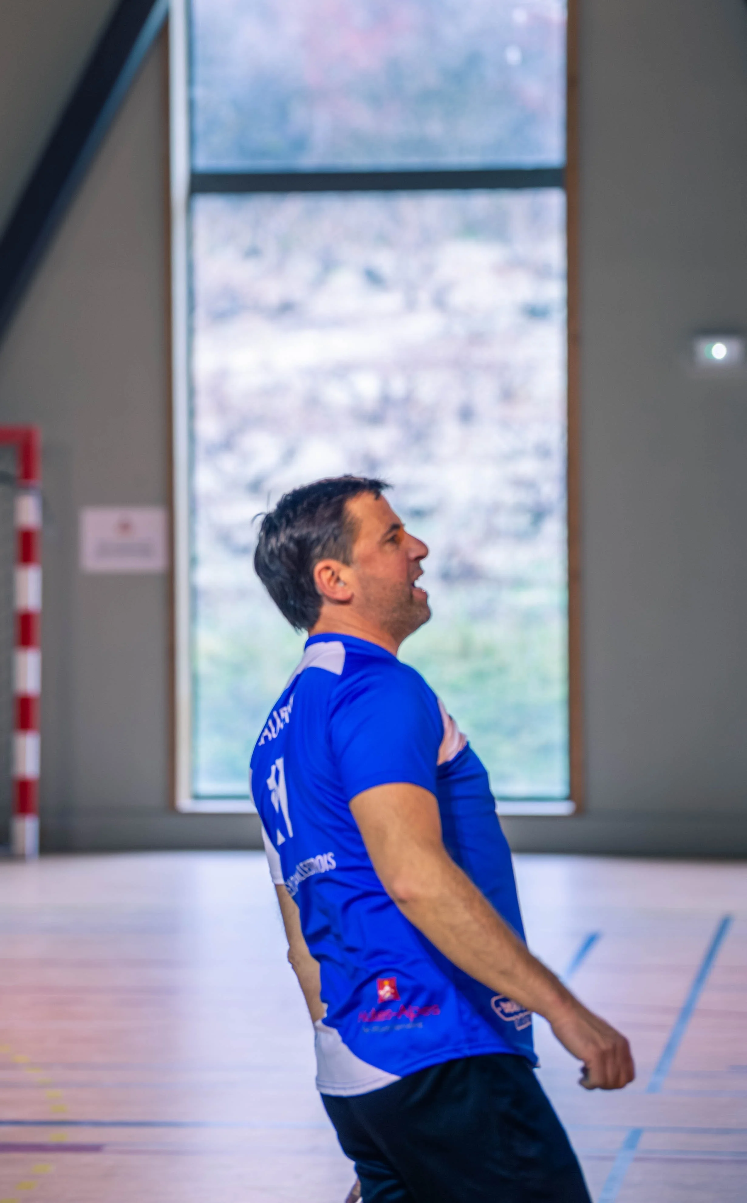 Un homme en maillot de sport bleu dans une salle de sport, faisant un geste de célébration ou de victoire, avec une grande fenêtre en arrière-plan.