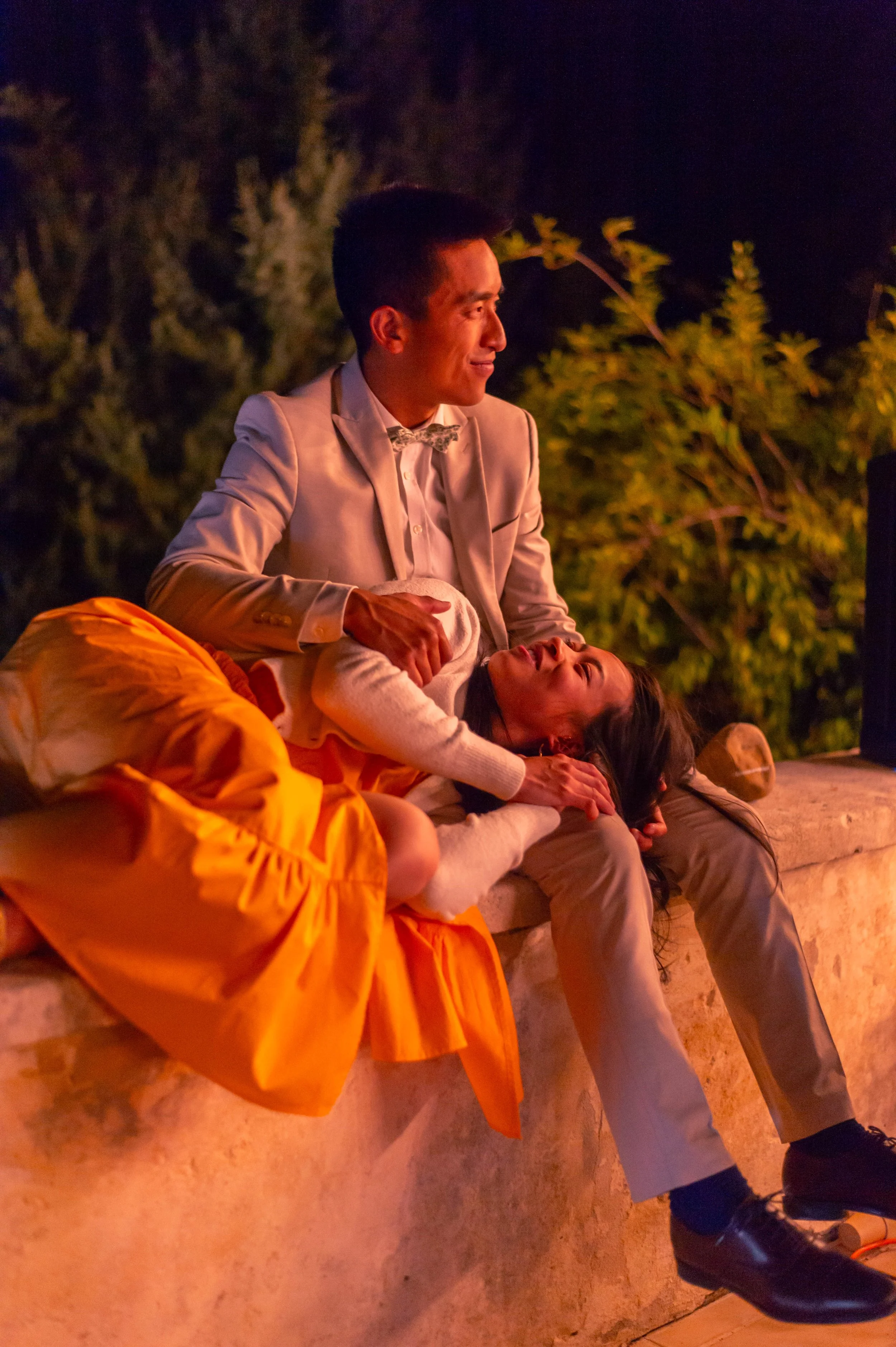 Un homme en costume beige et une femme en robe orange sont assis sur un mur en pierre, avec la femme allongée sur la personne en costume, dans un décor nocturne avec des arbres en arrière-plan.