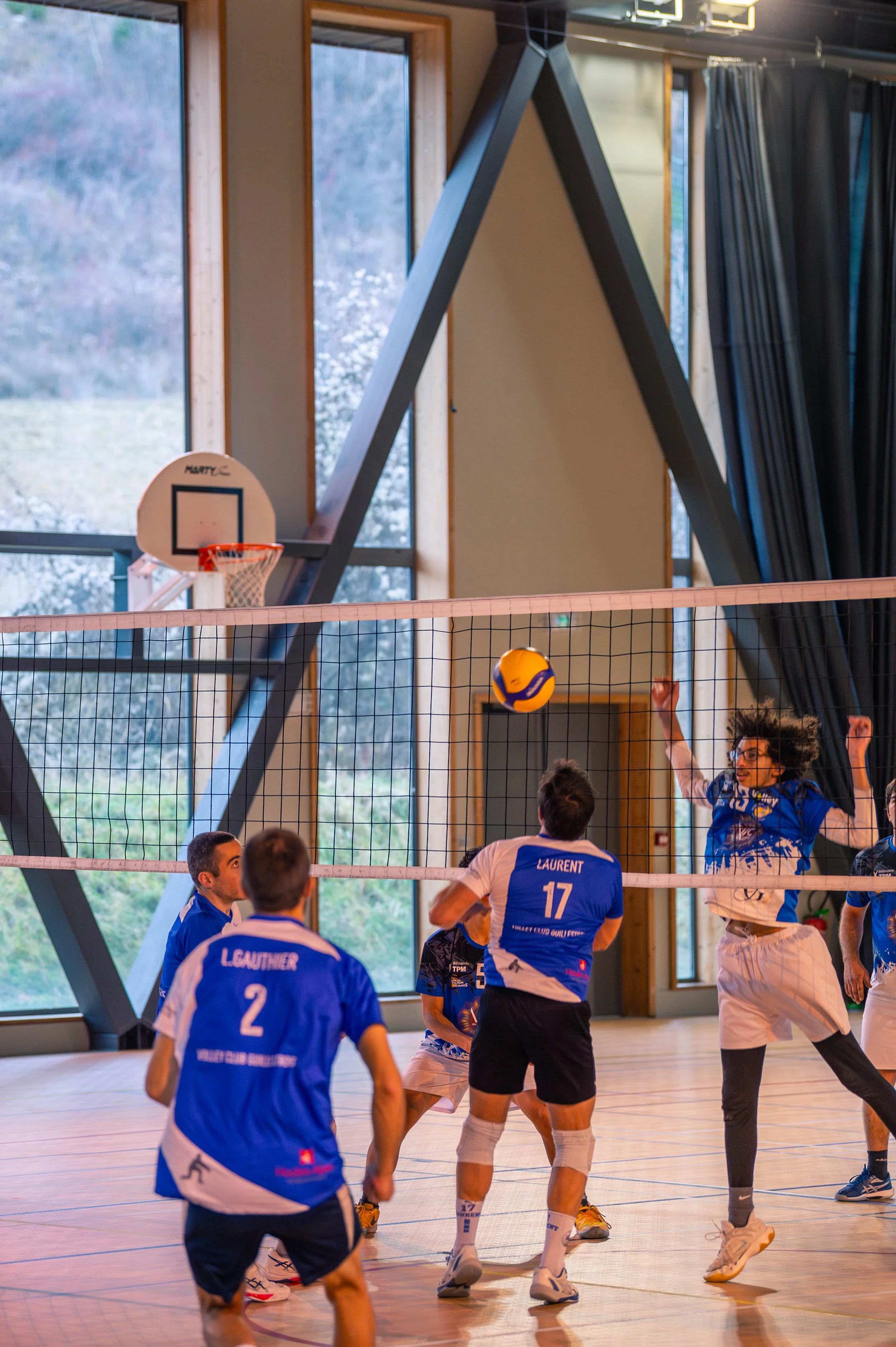 Joueurs de volley-ball en pleine action à l'intérieur d'un gymnase, avec un ballon au-dessus du filet, et un tableau de basketball en arrière-plan.