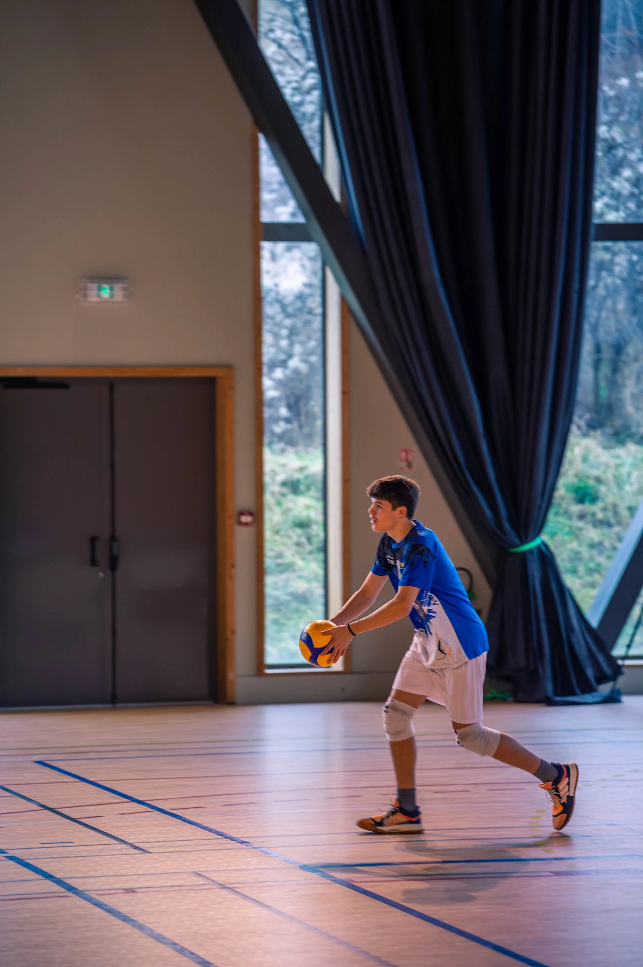 Jeune garçon jouant au volley-ball en intérieur, tenant un ballon, dans une salle aux grandes fenêtres et rideaux noirs.