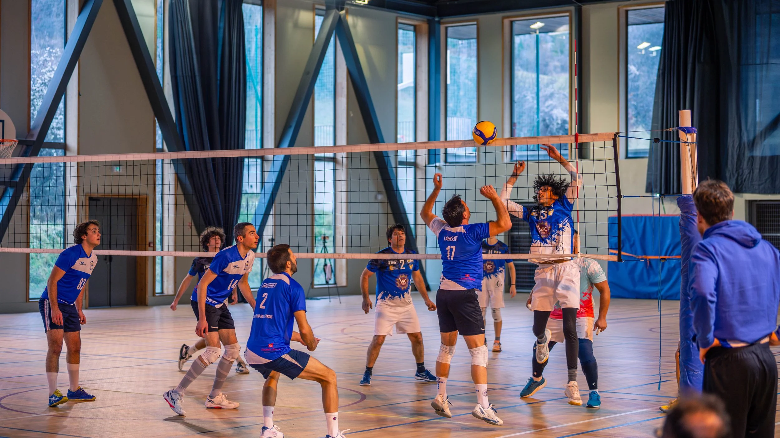 Un groupe de joueurs de volley-ball en action à l'intérieur d'une salle de sport avec un filet et plusieurs fenêtres.