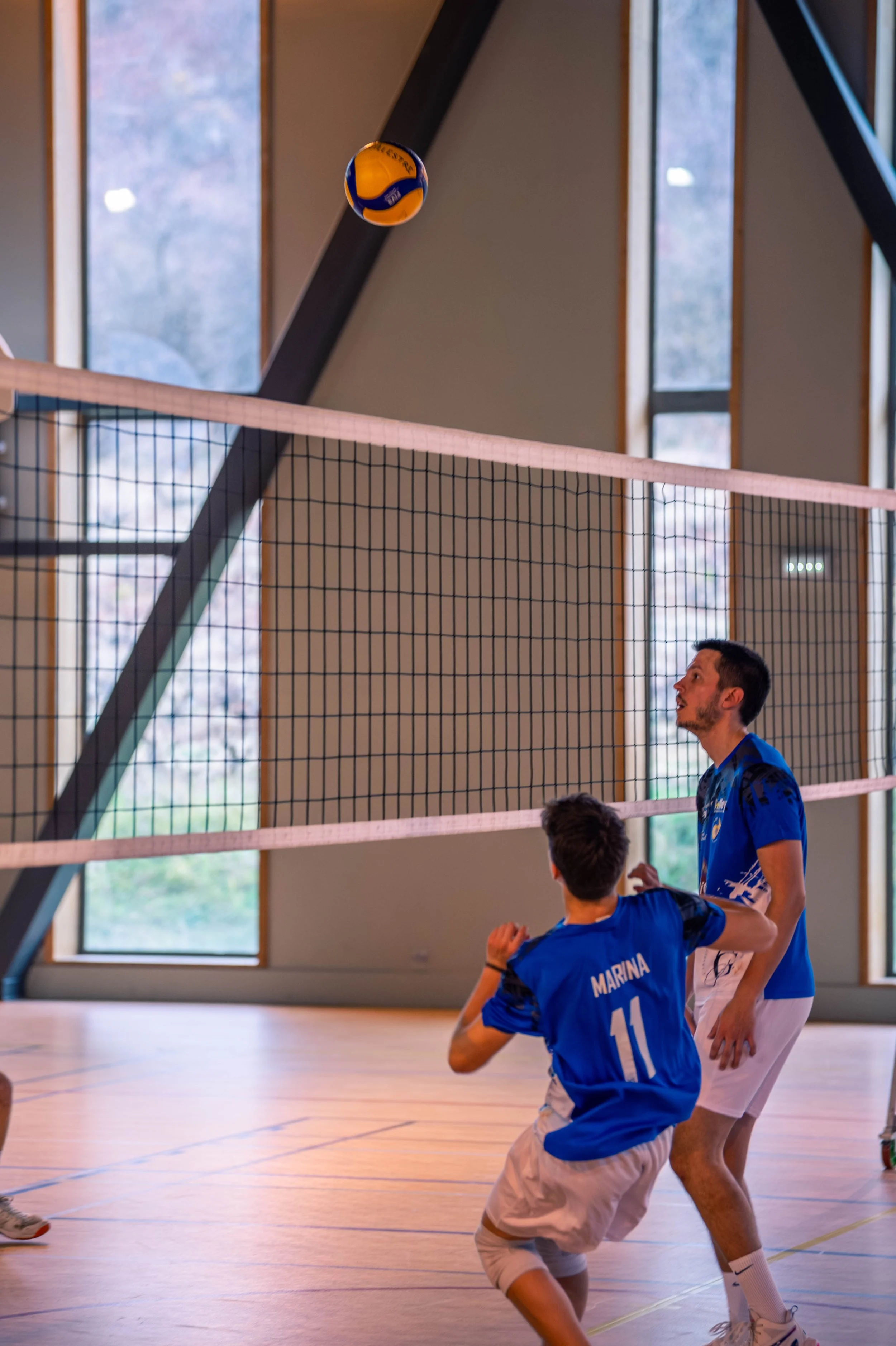 Deux joueurs en maillot bleu jouent au volleyball en intérieur, un préparant à recevoir la balle qui vole dans l'air.