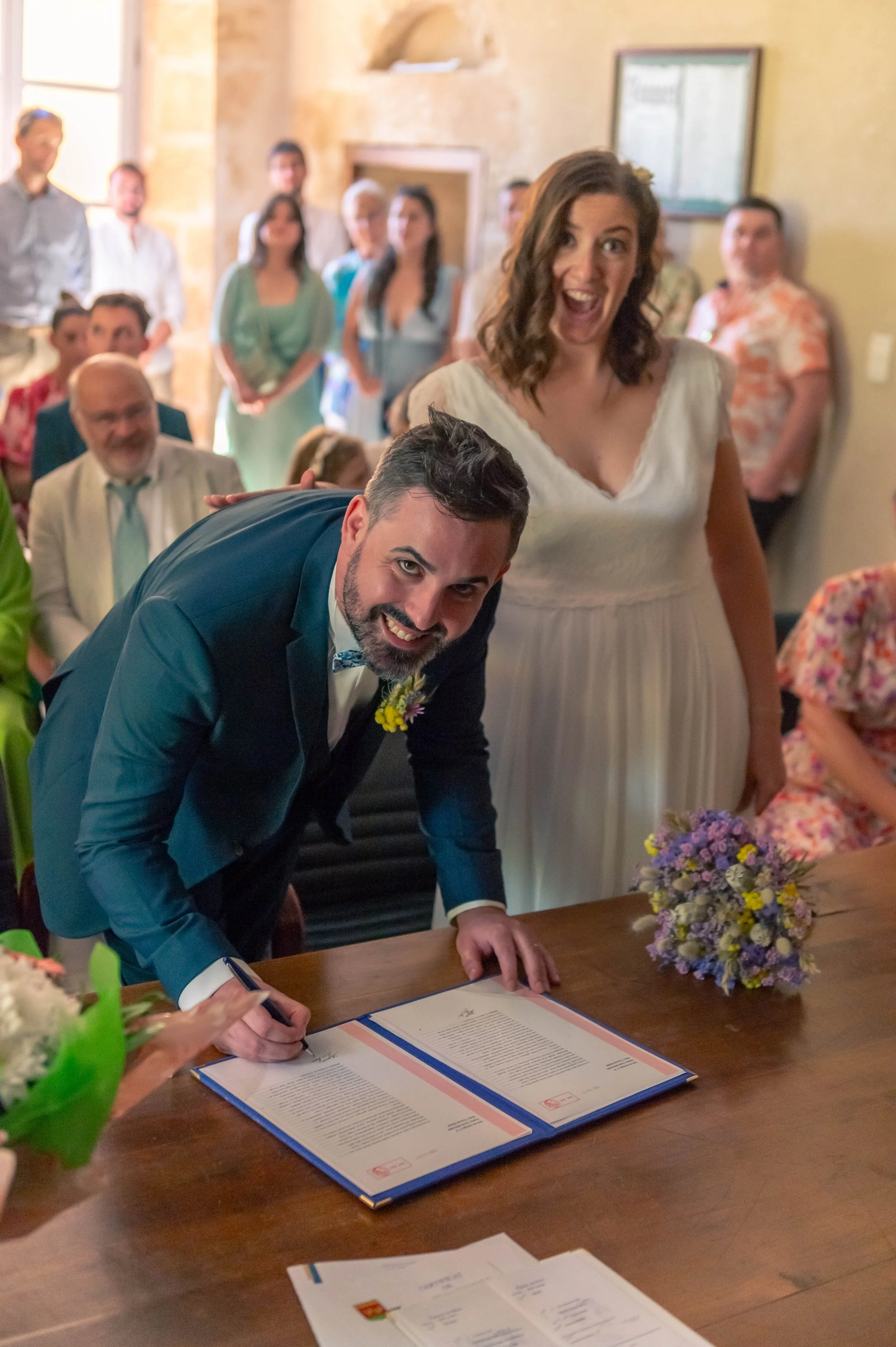 Un homme signe un document lors d'un mariage, entouré de guests souriants, une femme en robe blanche à ses côtés. L'ambiance est joyeuse dans une salle décorée.