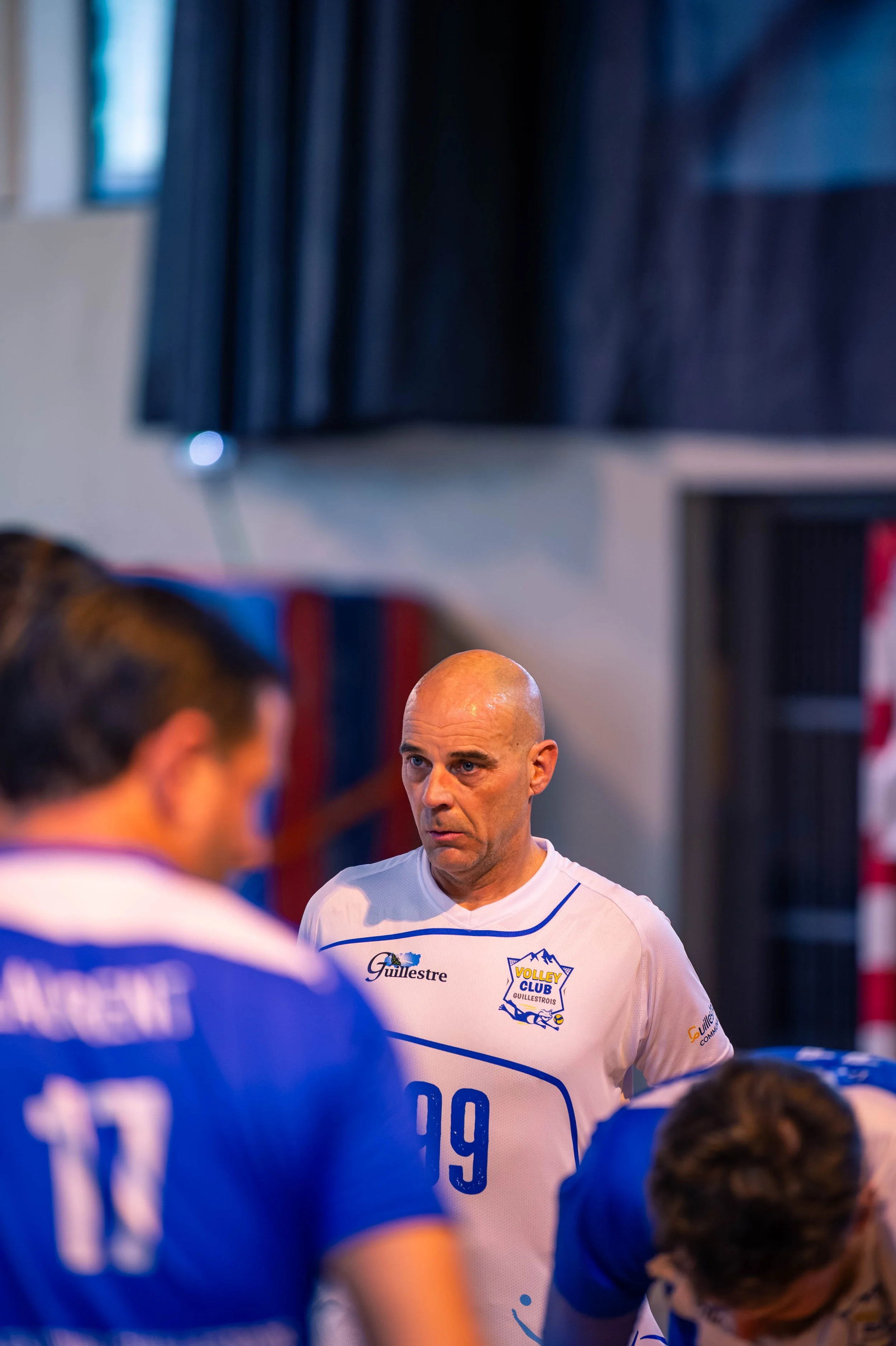 Un entraîneur de volleyball parle à ses joueurs durant un match, portant un maillot blanc avec le logo du club de volley Guilesttre.