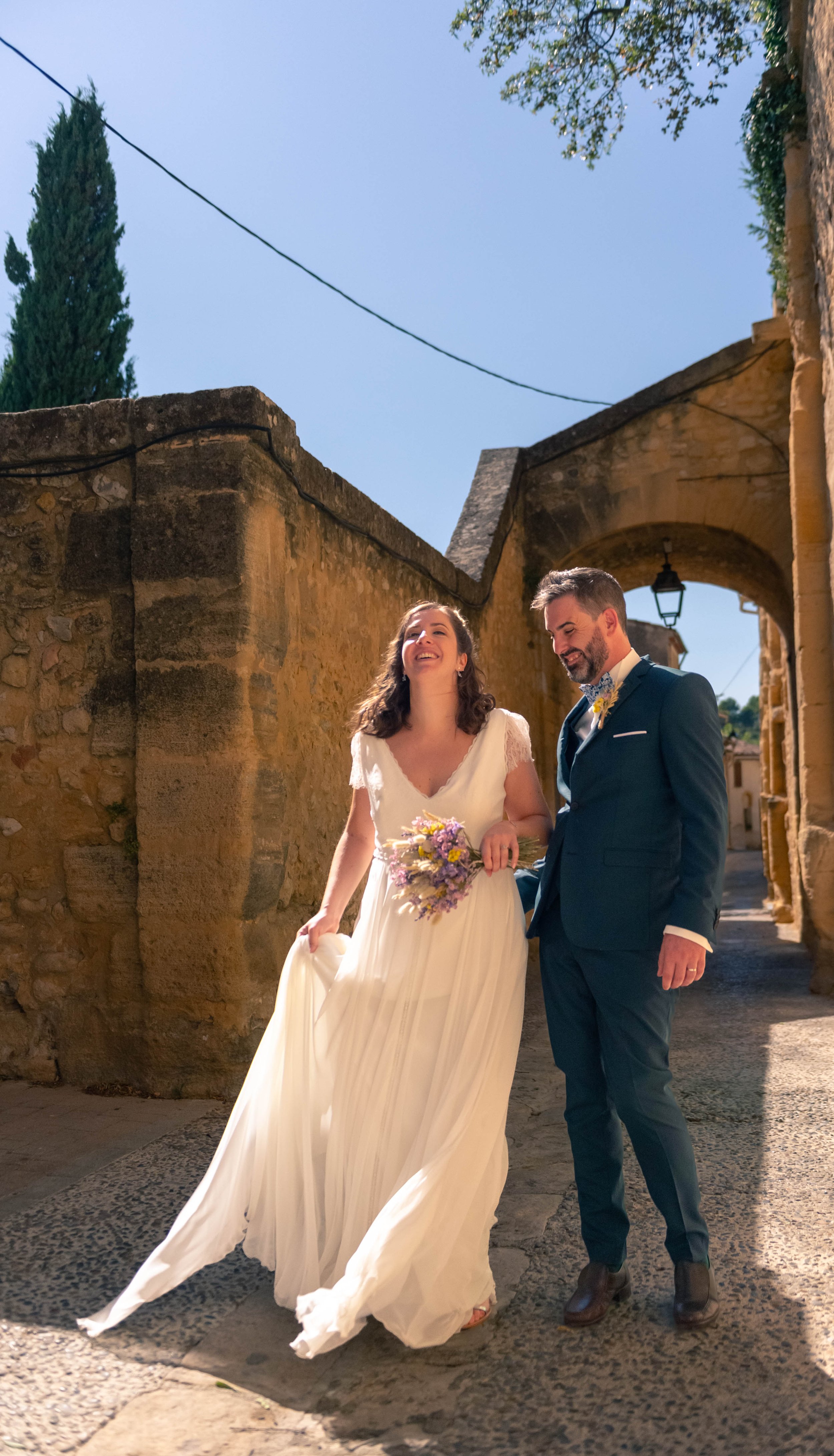 Un couple de mariés marche dans une rue pavée, la mariée porte une robe blanche et tient un bouquet, le marié porte un costume sombre, ils sourient et semblent heureux.