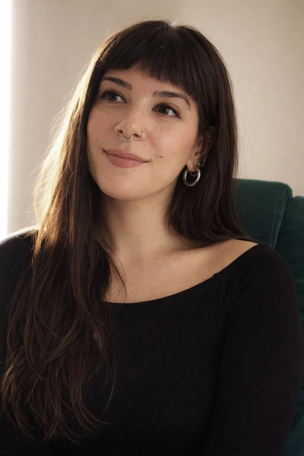 A woman with long brown hair and bangs, wearing a septum ring and hoop earrings, sitting in a green chair, wearing a black top.
