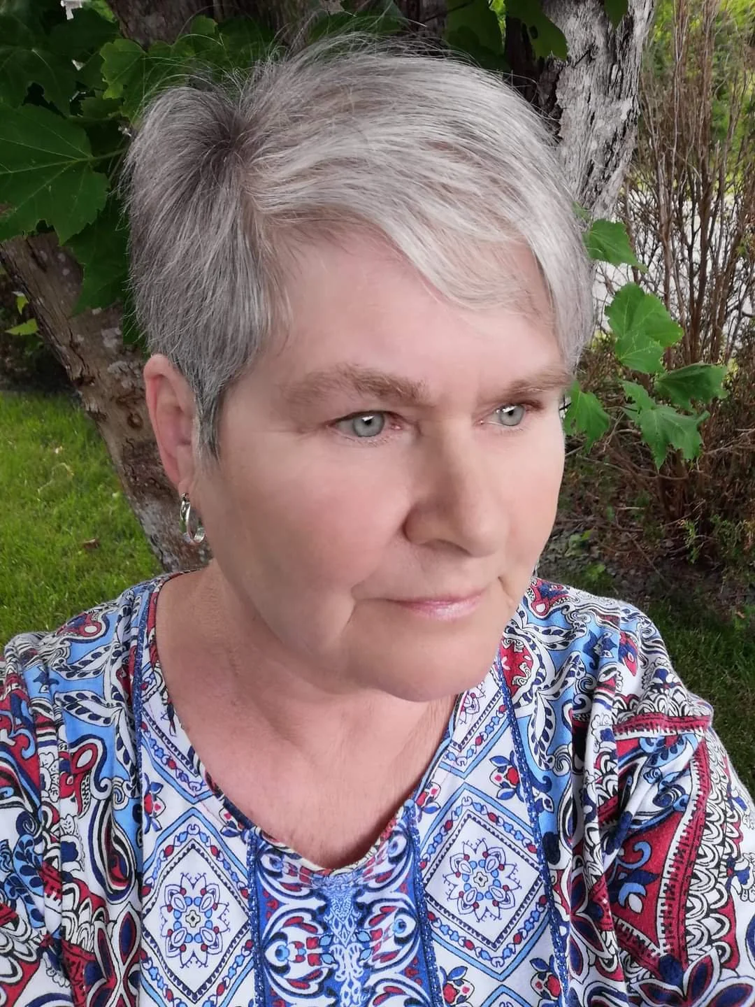 Close-up of a woman with short gray hair and blue eyes, wearing earrings and a colorful patterned top, outdoors near a tree with green leaves.