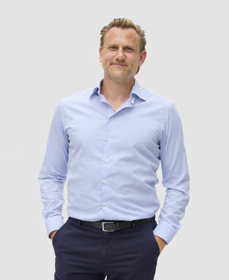 A middle-aged man with short light brown hair and a beard wearing a light blue button-up shirt and dark pants, standing against a plain white background.