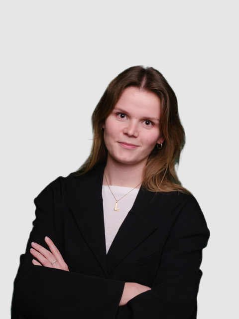 A young woman with shoulder-length brown hair wearing a black blazer and a white shirt, crossing her arms and smiling slightly against a plain light gray background.