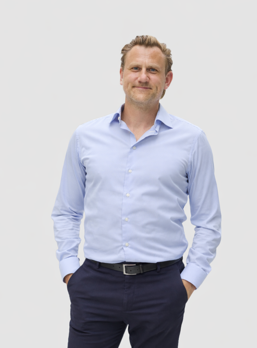 Man in light blue dress shirt and dark pants standing with hands in pockets against a plain white background.