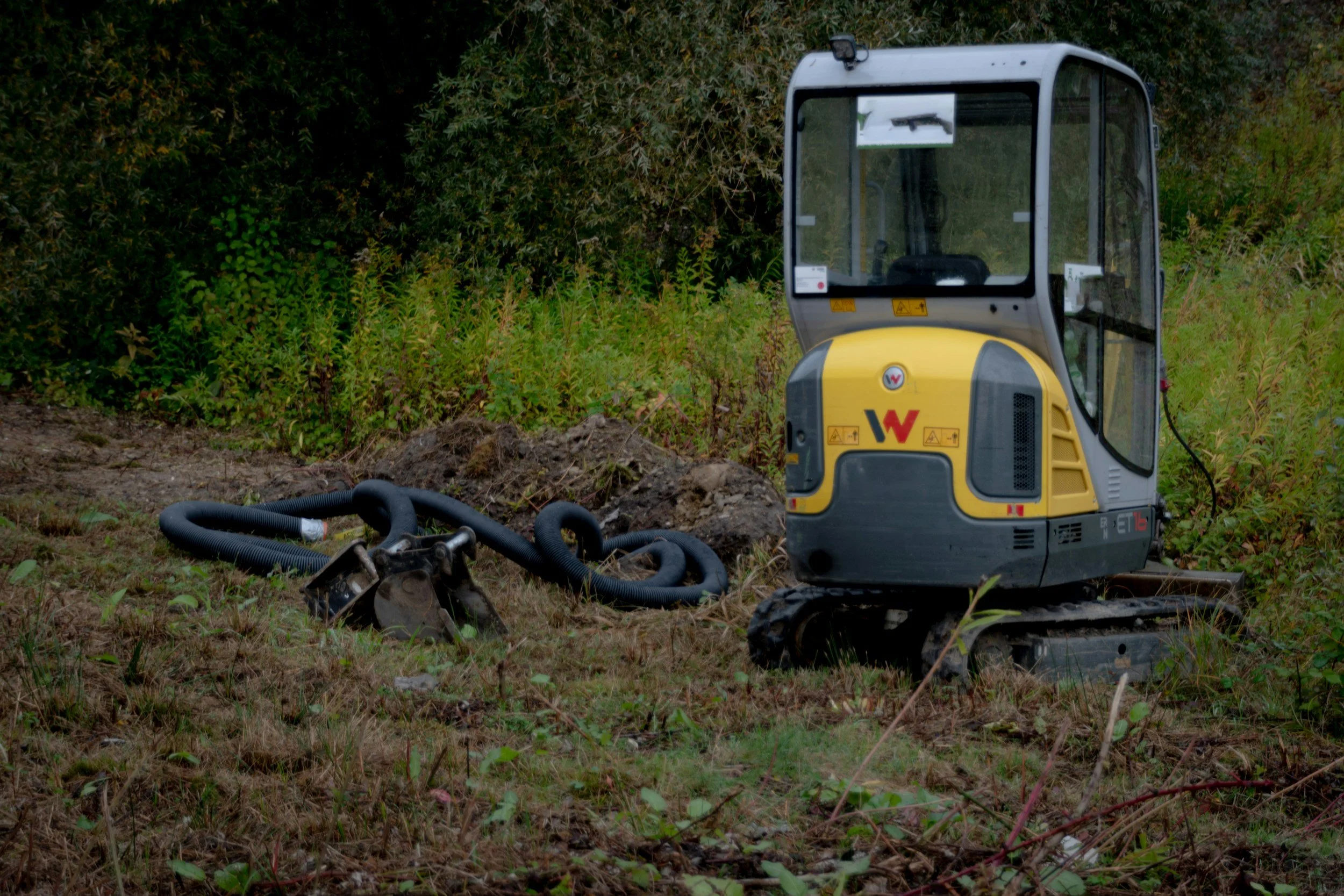 A small tracked excavator with a yellow and gray body sitting on grass with a black hose nearby, surrounded by greenery.