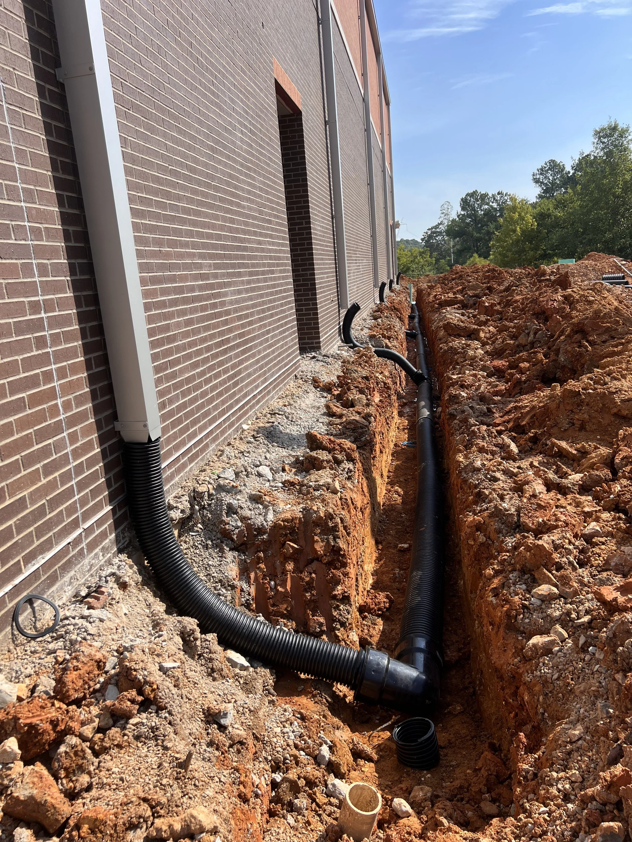 Construction site showing trench with black drainage pipes alongside a brick building, with trees and blue sky in the background.