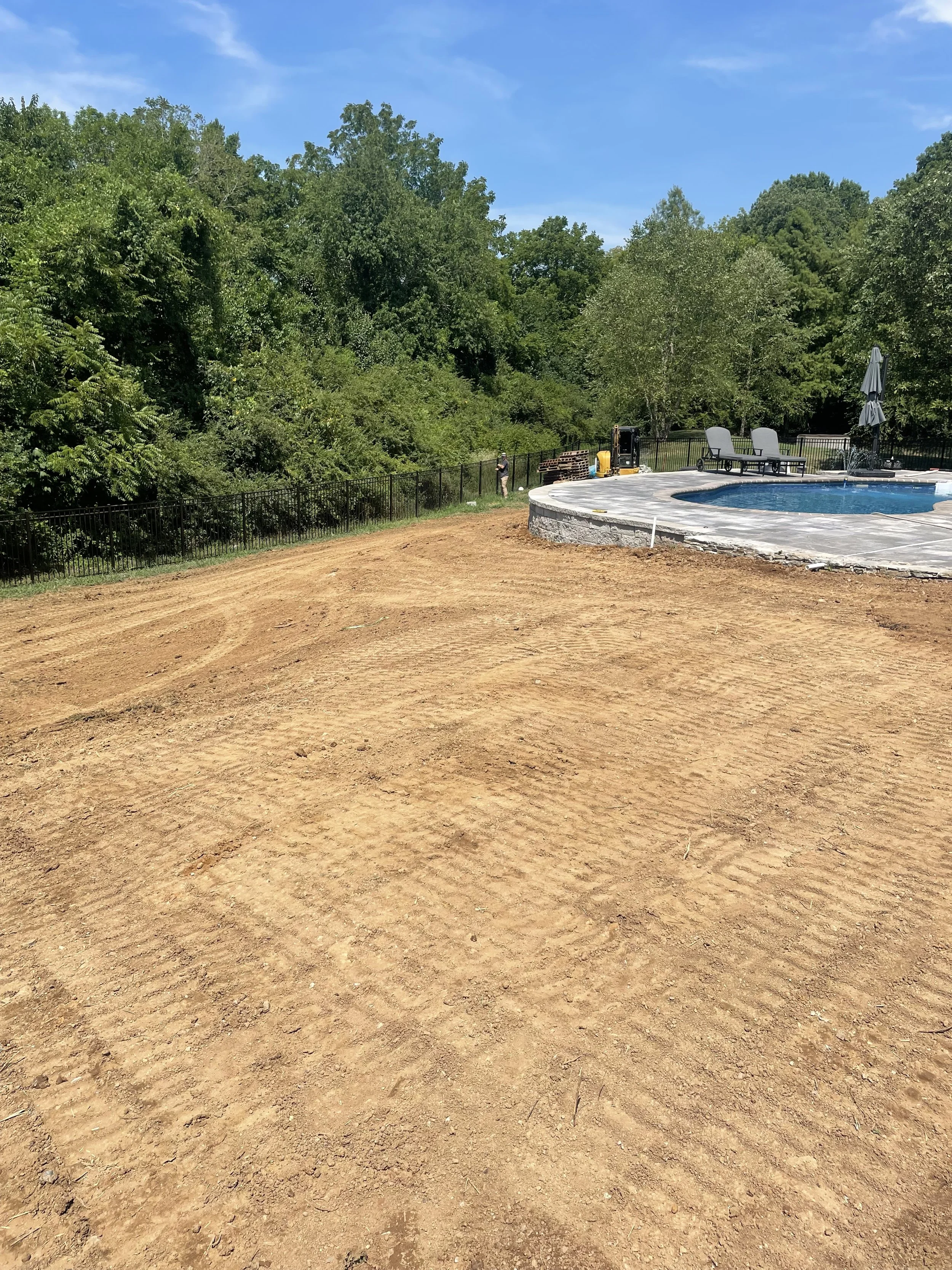 Construction area with dirt ground next to a partially built swimming pool, surrounded by trees and a black metal fence.