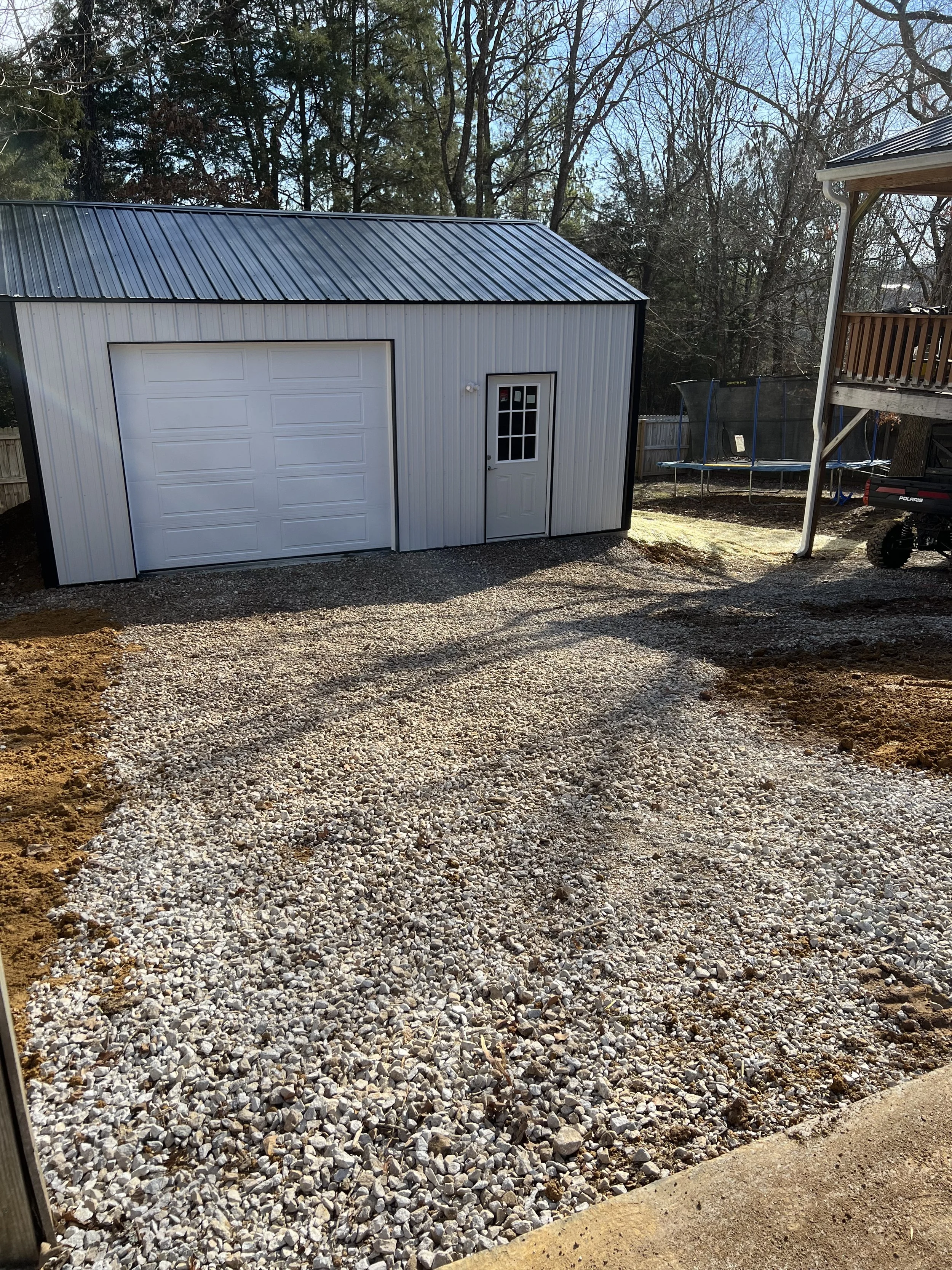 A newly laid gravel driveway leading to a white metal storage shed with a garage door and a small door with window panes, located in a backyard with trees, a trampoline, a deck, and some equipment.