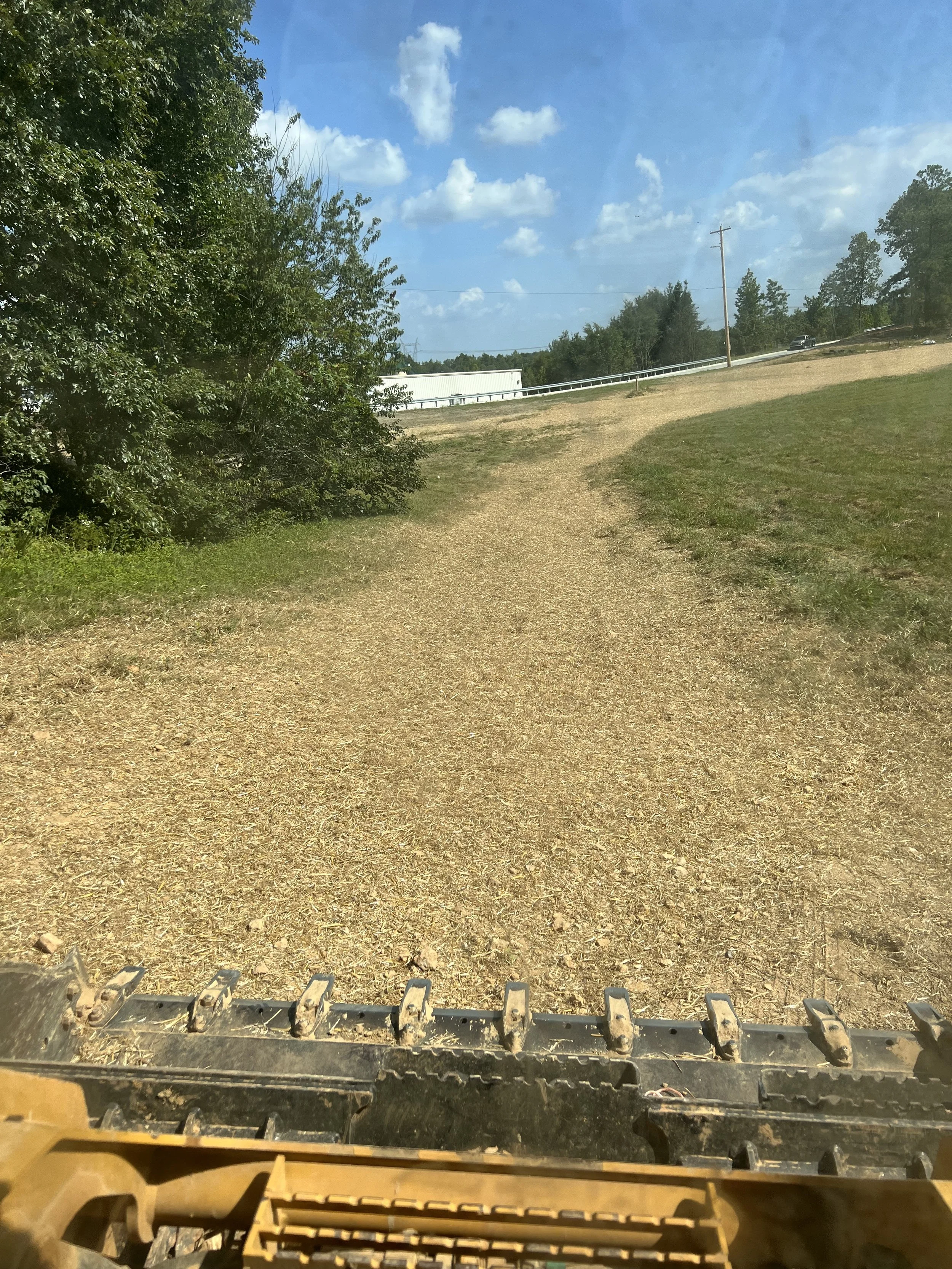View from a vehicle's front attachment showing a dirt path through a grassy area, trees on the left, a white building, and a blue sky with clouds in the distance.