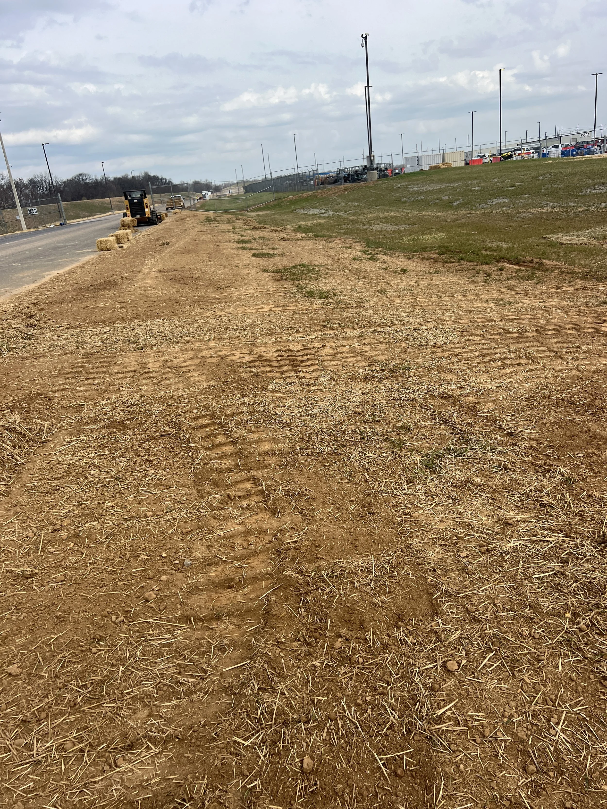 A dirt path next to a paved road with hay bales and a small vehicle parked along the road. Construction fencing and parked cars are visible in the background under a cloudy sky.