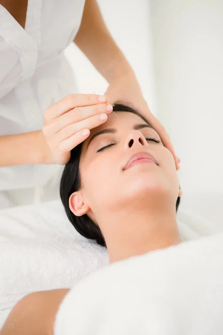 A woman receiving a facial massage or skincare treatment from a professional at a spa or clinic, lying with her eyes closed and smiling.