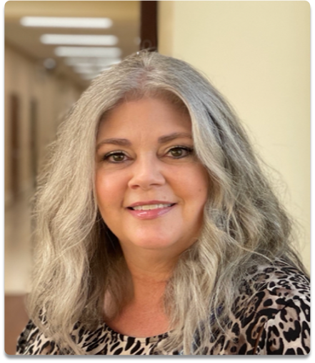 A woman with long gray hair smiling in a hallway.