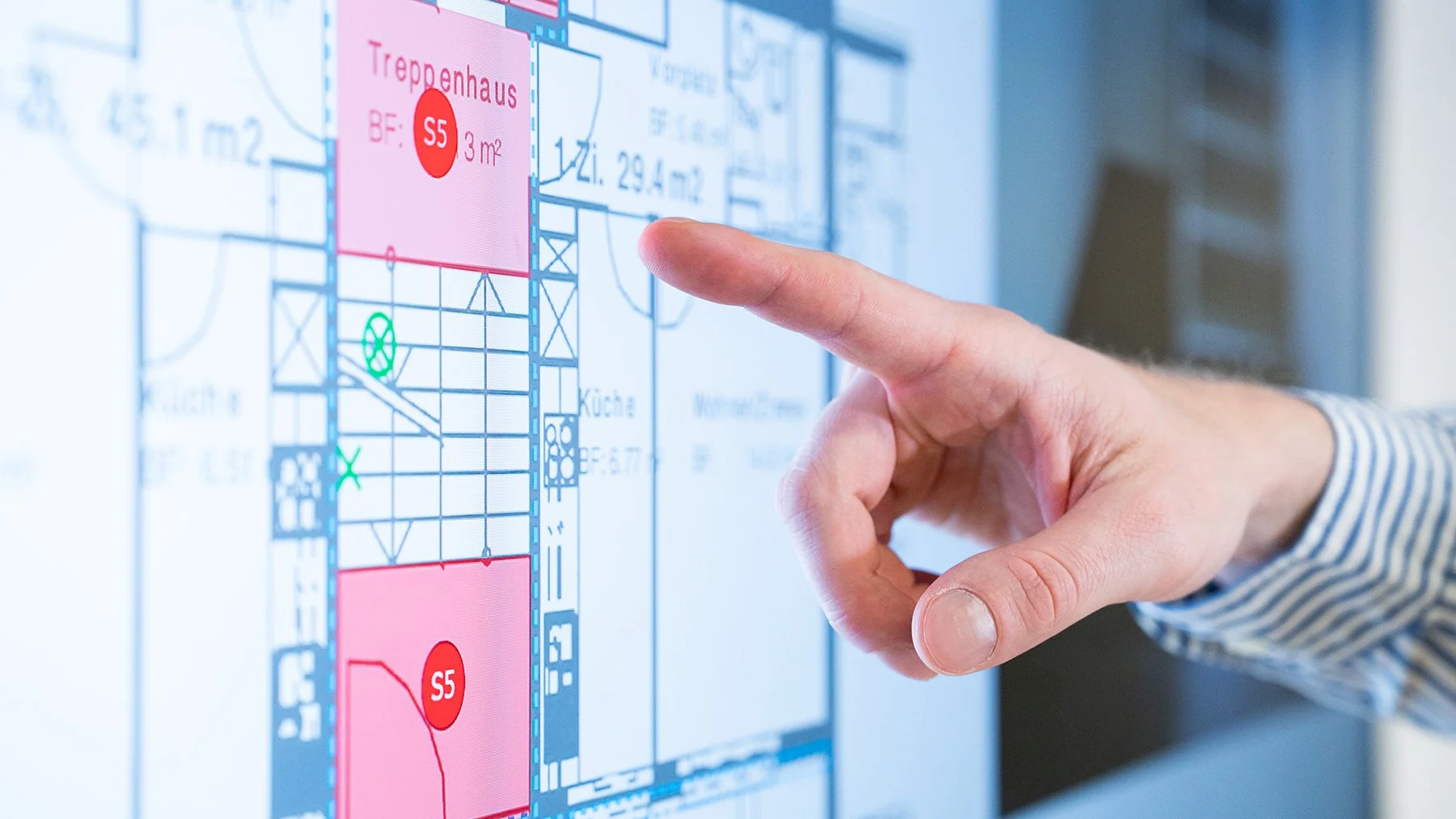 Person zeigt auf einen digitalen Grundrissplan auf einem Bildschirm, der Zimmer und Flächen mit Beschriftungen wie 'Treppenhaus' und 'Küche' enthält.
