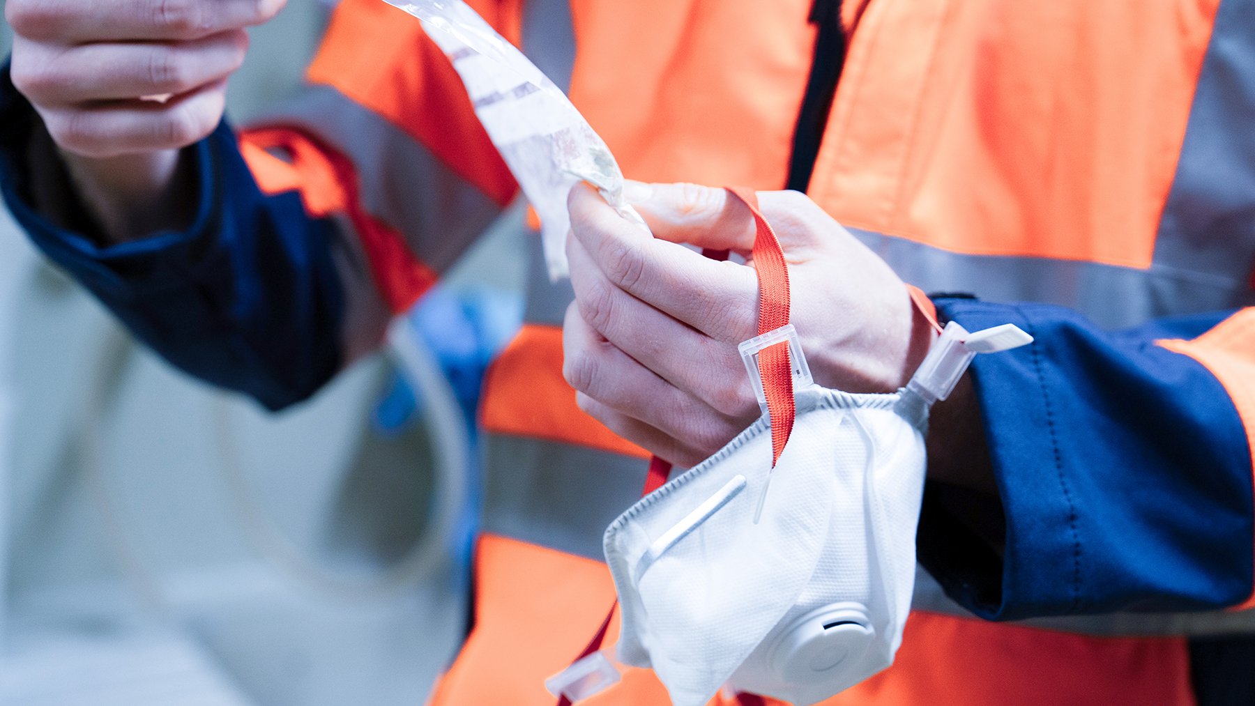 Person in Schutzkleidung legt eine Schutzmaske mit Atemschutzfilter in eine Stofftasche. Person trägt eine auffällige orange Sicherheitsweste.