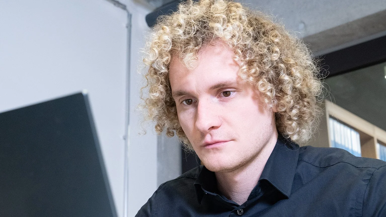 Junger Mann mit lockigem blondem Haar arbeitet am Laptop in einem modernen Büro.