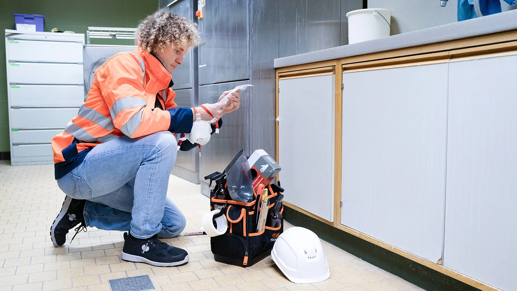 Bodenarbeiter sitzt auf dem Boden und liest einen Zettel, trägt Sicherheitsschuhe, orange Warnweste, in der Nähe Werkzeugsatz, Schutzhelm, in einem Raum mit grauen Schränken.