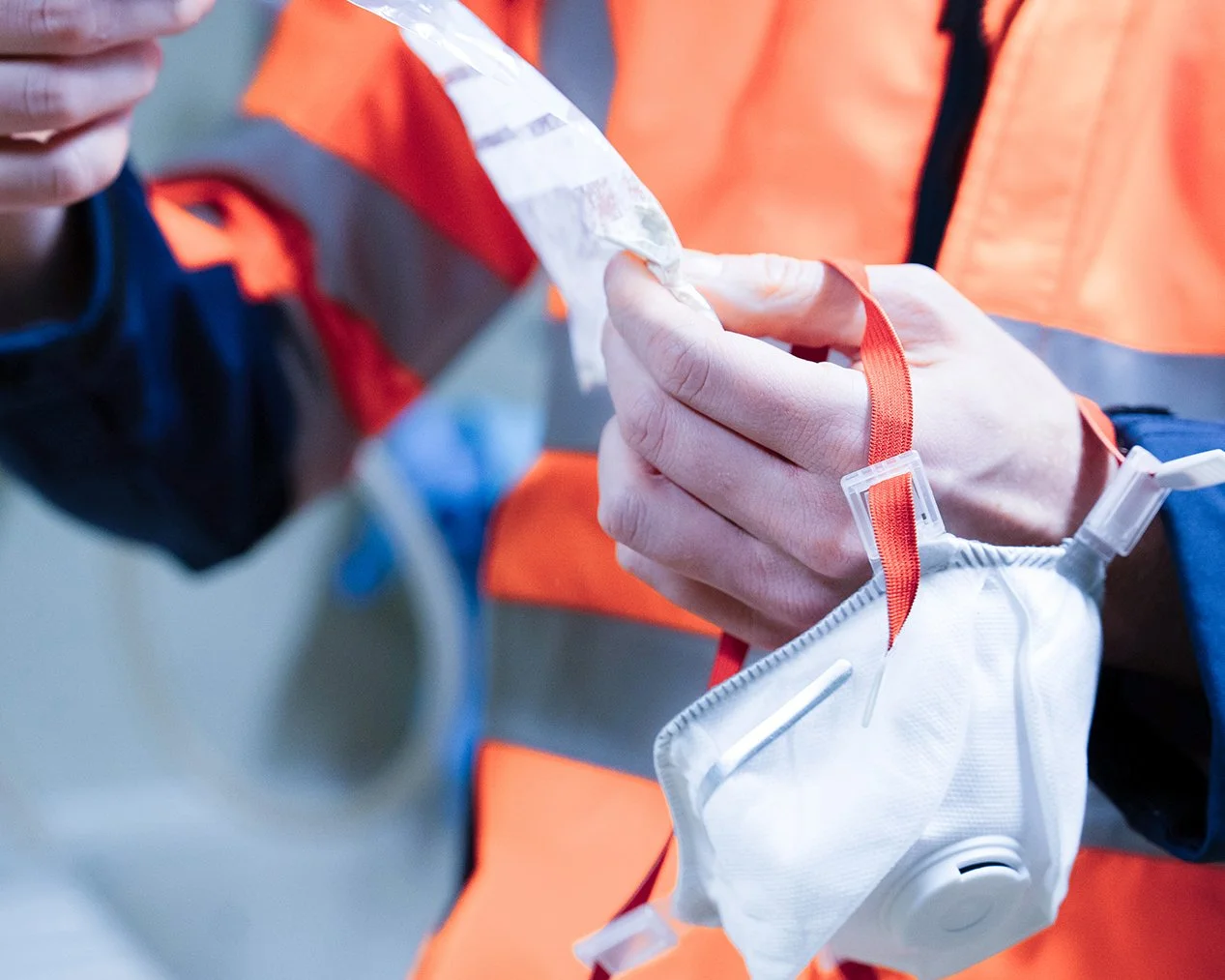 Eine Person in einem orangefarbenen Sicherheitsweste hält eine medizinische Maske mit einem Filter in den Händen.