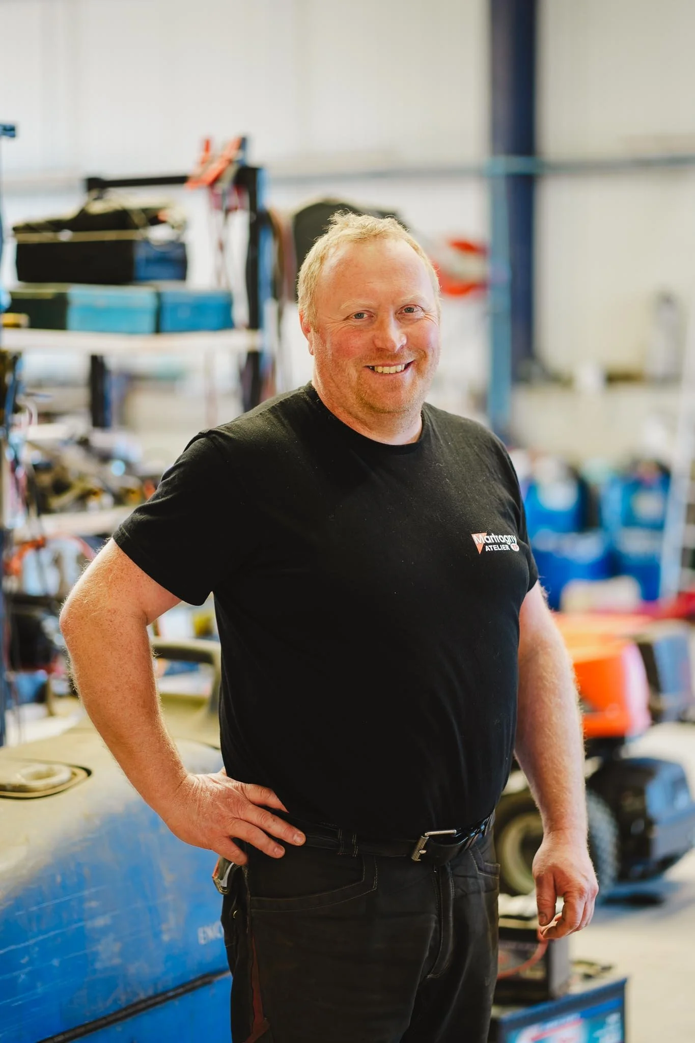 Un homme souriant dans un atelier de réparation ou de maintenance, entouré d'outils et d'équipements.