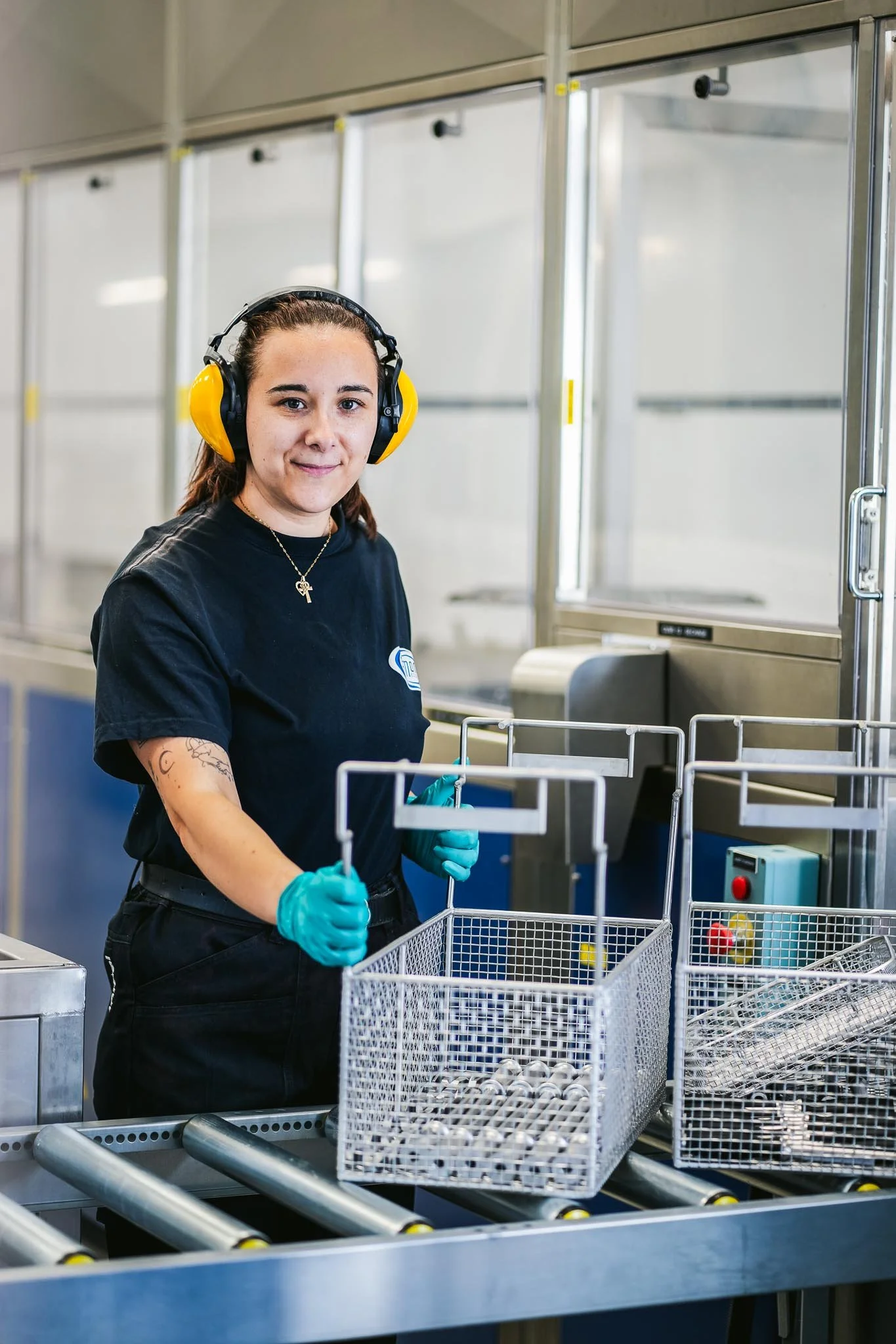 Une femme portant des protections auditives, des gants en caoutchouc, et un t-shirt noir, travaille dans une usine ou une salle de confection avec des cages de pièces métalliques sur un convoyeur.
