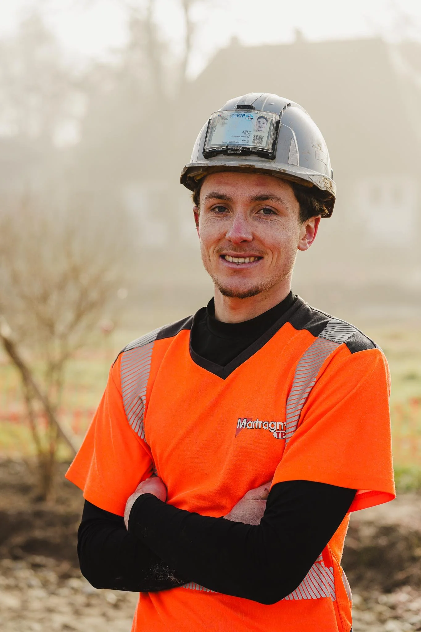 Un jeune homme en tenue de travail orange portant un casque de sécurité et un t-shirt avec le nom 'Martragny' pose avec les bras croisés dans un extérieur par temps ensoleillé.