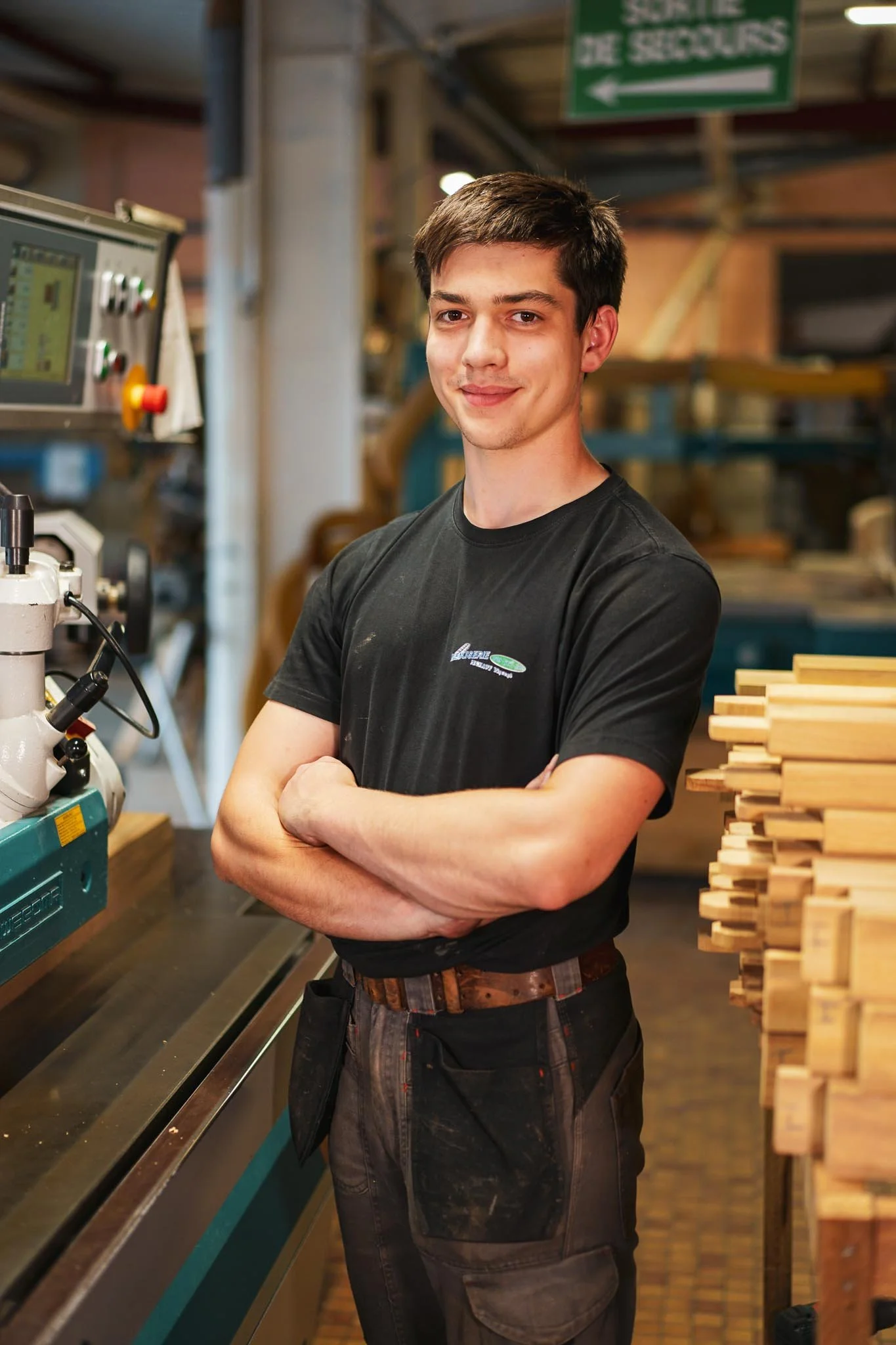Un jeune homme en vêtements de travail dans un atelier de menuiserie, tenant ses bras croisés et souriant, entouré de machines et de morceaux de bois.