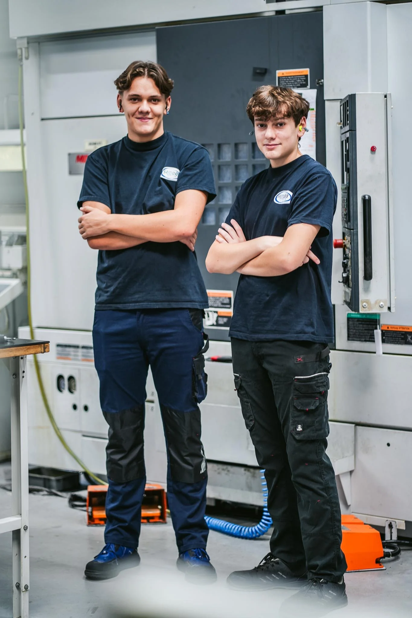 Deux jeunes hommes portent des uniformes de travail dans un atelier industriel, debout côte à côte avec les bras croisés, devant des machines et équipements techniques.