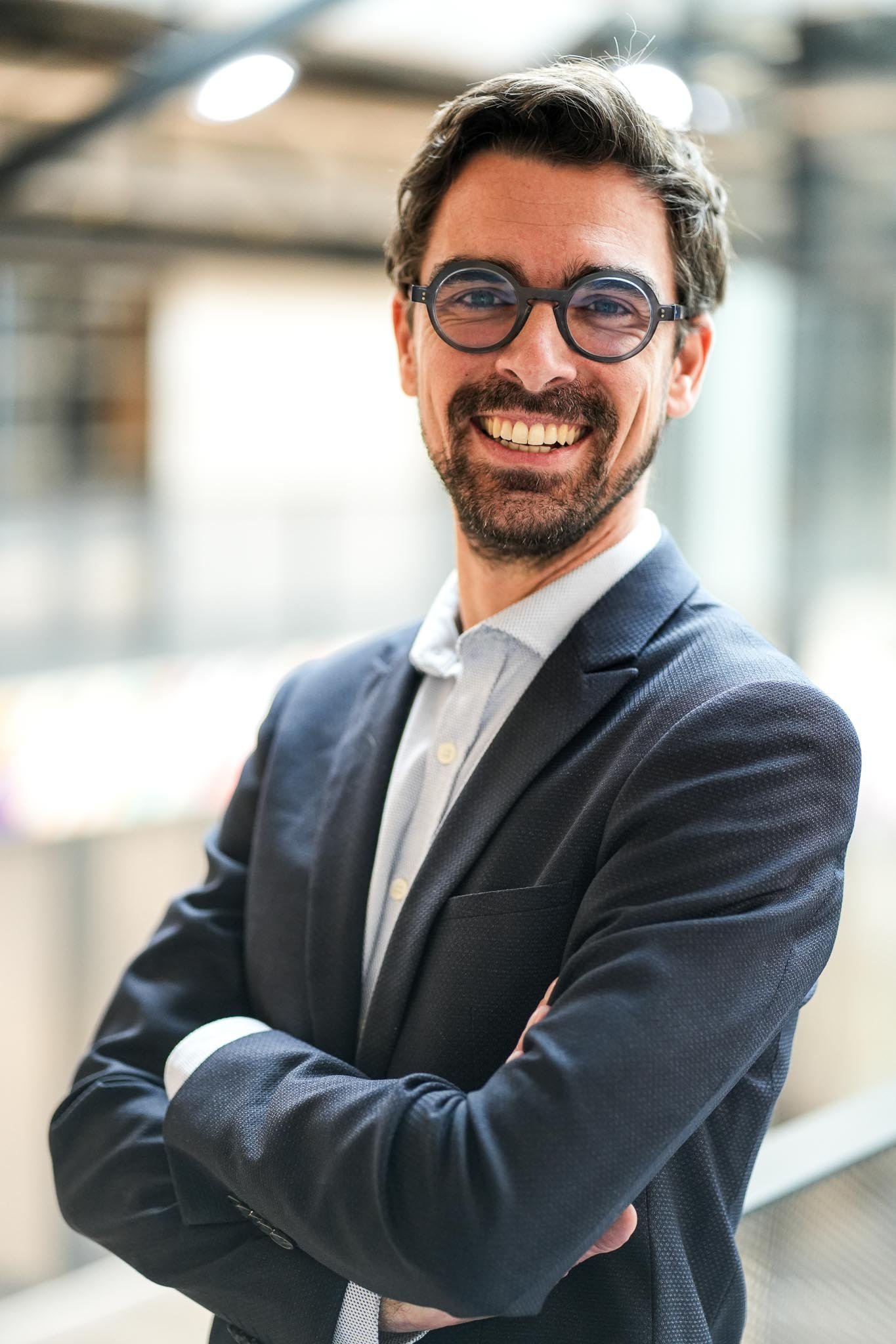 Homme d'affaires souriant portant des lunettes rondes, en costume cravate, avec les bras croisés dans un espace moderne et lumineux.