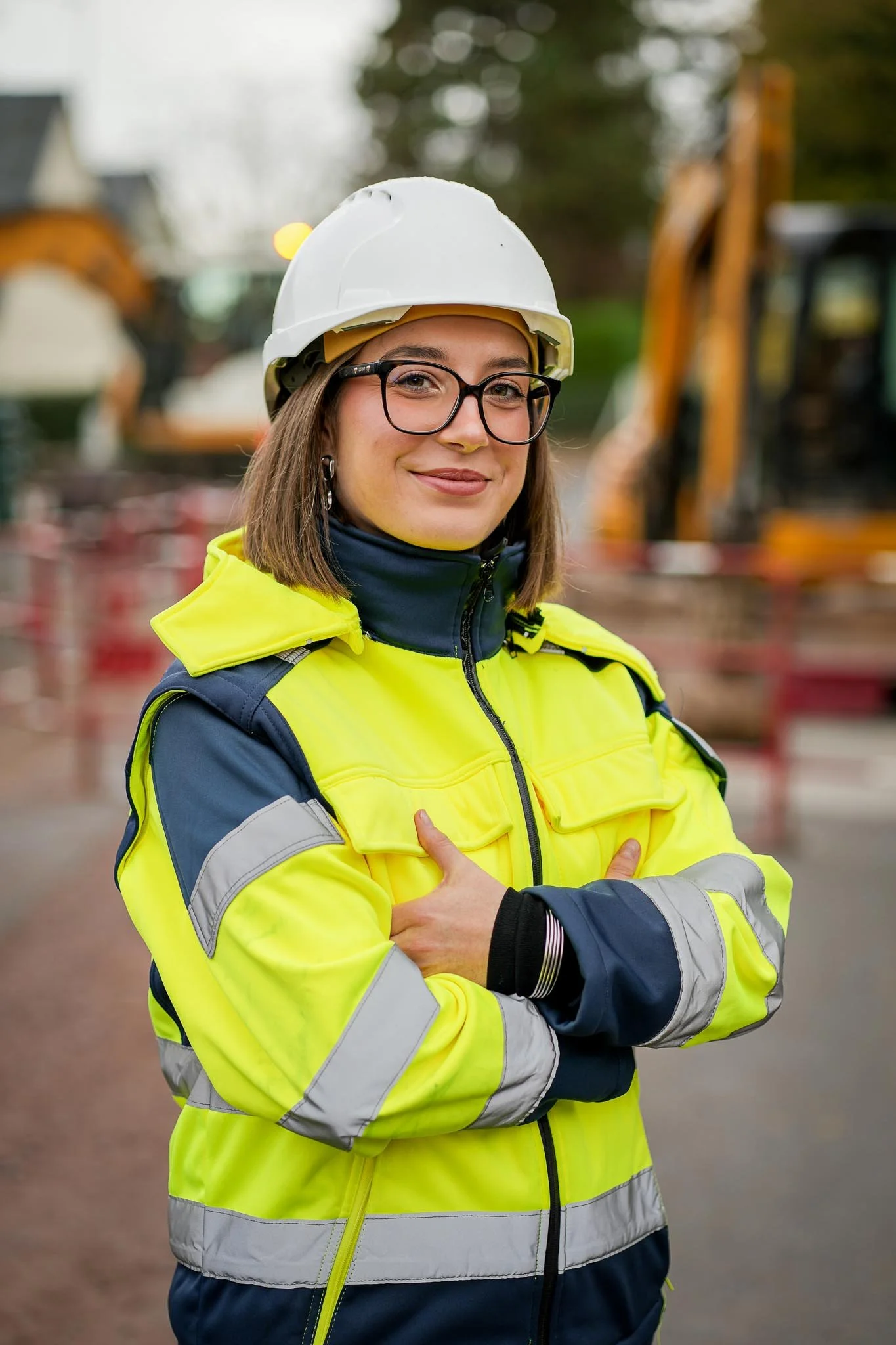 Une femme portant un casque blanc, des lunettes et une veste de sécurité jaune fluo avec des détails gris et bleus. Elle se tient avec ses bras croisés devant un chantier.
