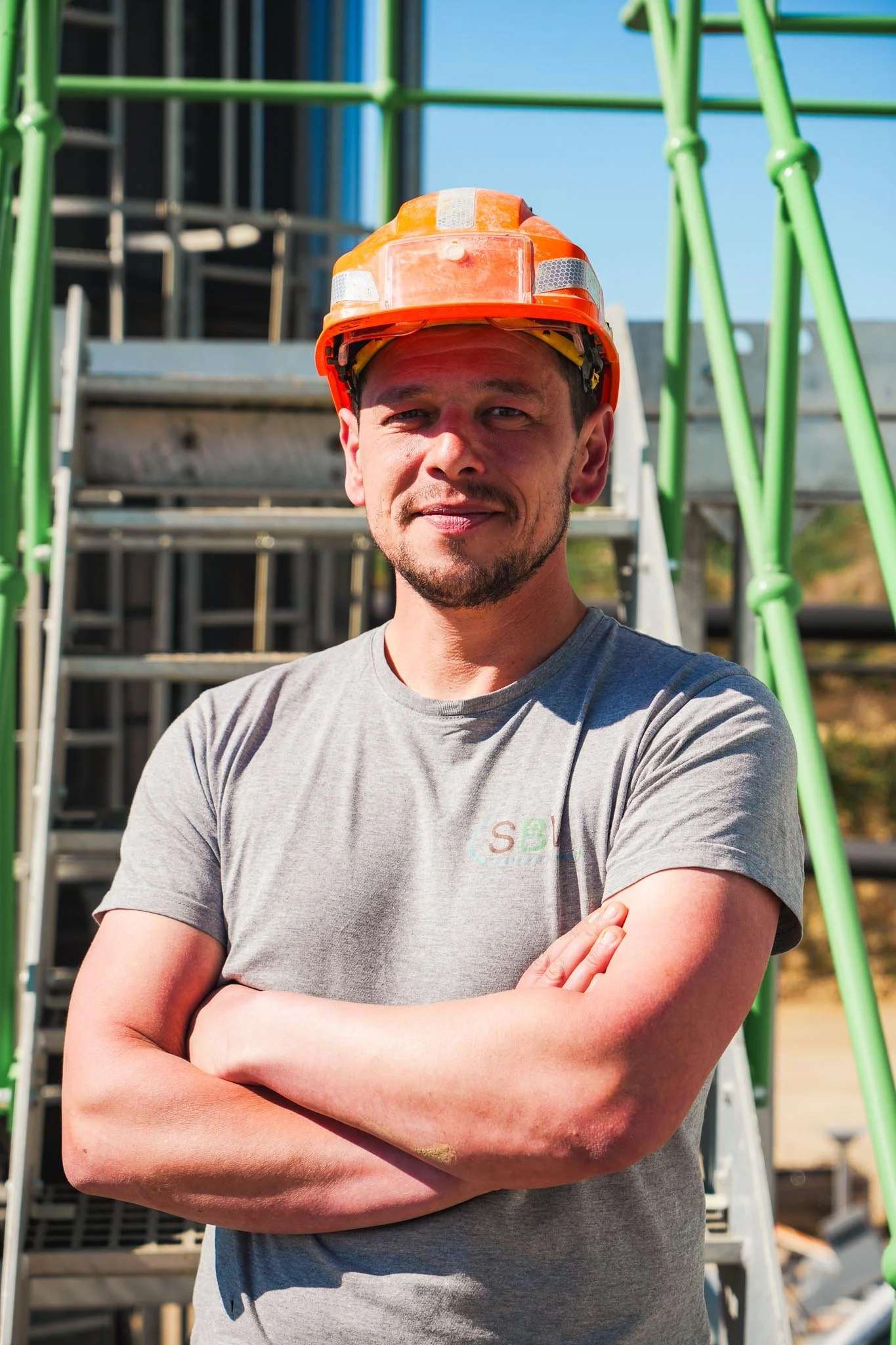 Un homme avec un casque orange se tient devant une structure d'échafaudage verte, les bras croisés, sous un ciel bleu clair.