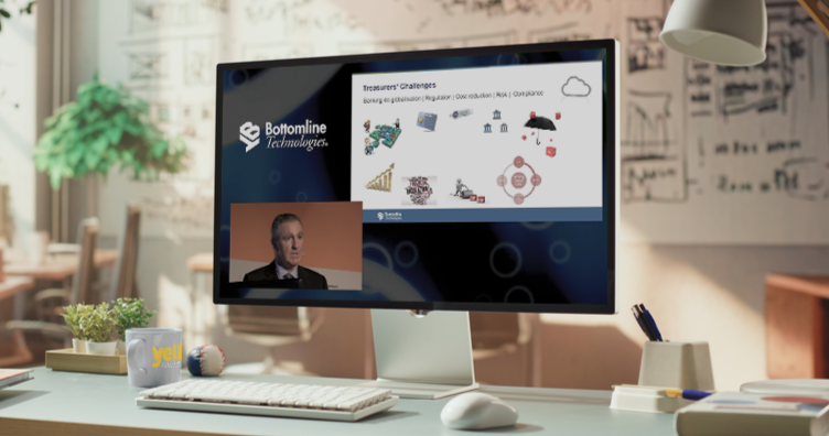 A computer monitor displaying a virtual presentation with the logo 'Bottomline Technologies' and a speaker in a small window. The presentation slide covers treasury challenges with icons and charts. The desk has a mug, a ball, and office supplies, and the background shows a well-lit office with bookshelves and artwork.