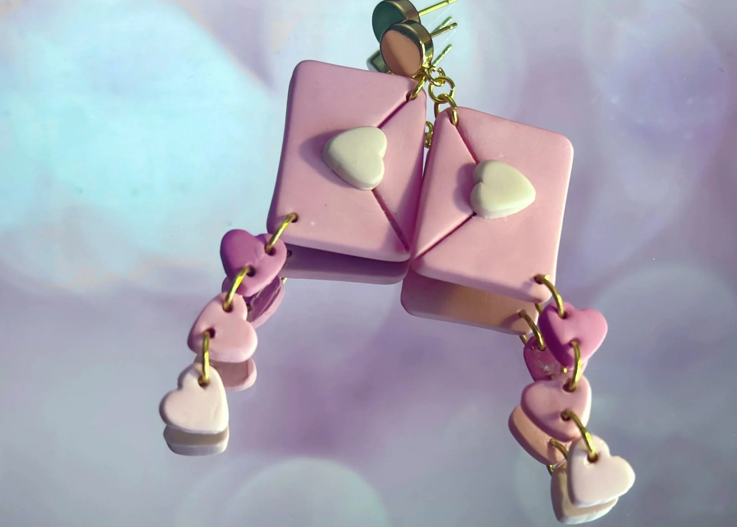 Decorative pink and lavender heart-shaped and rectangular beads on a gold chain, with small white heart accents, hanging against a soft, iridescent background.