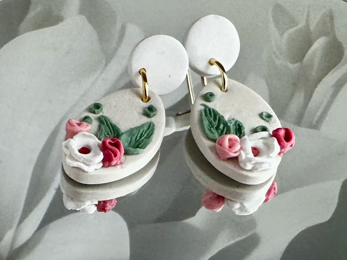 White floral earrings with pastel pink and green leaf accents, hanging on a white background.