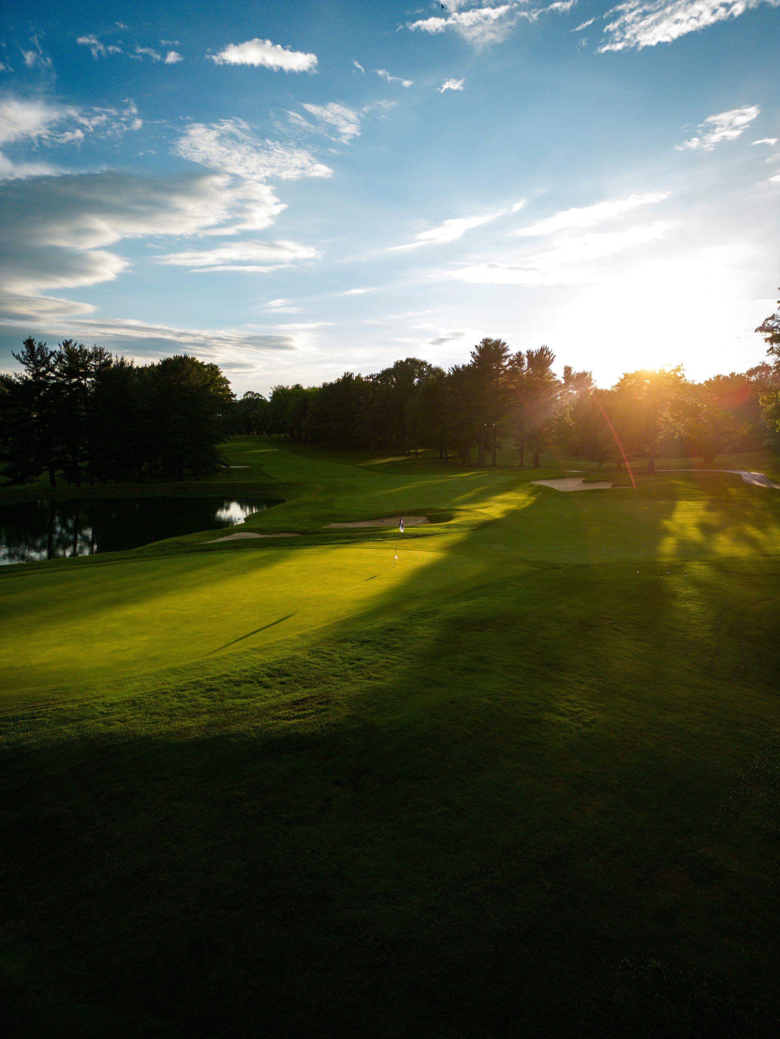 A scenic golf course at sunset with lush green fairways, a small pond on the left, and a flag on the green. Tall trees line the course, and the sun is setting behind the horizon, casting warm light and shadows across the landscape.