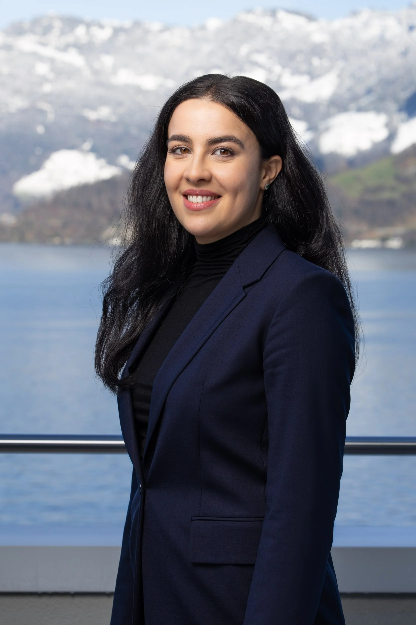 Frau mit schwarzem Haar und dunklem Blazer vor Berglandschaft mit Schnee und See.
