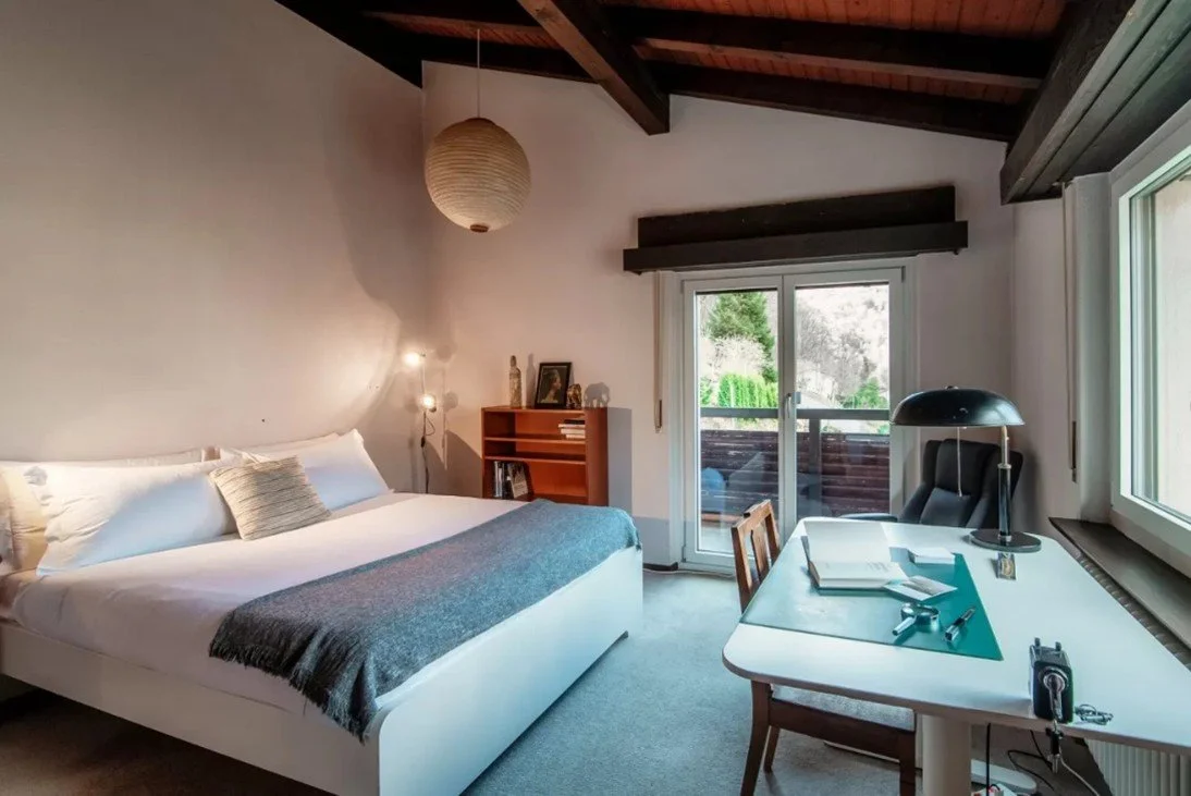 Ein Schlafzimmer mit einem Doppelbett, einem Schreibtisch mit Unterlagen und Stauraum, einem Fenster und einer Terrasse im Hintergrund.